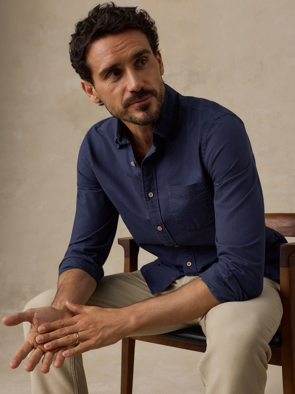 A man wearing a navy blue long-sleeve button-down shirt and beige trousers, sitting on a wooden chair against a textured beige wall., image 2 of 9