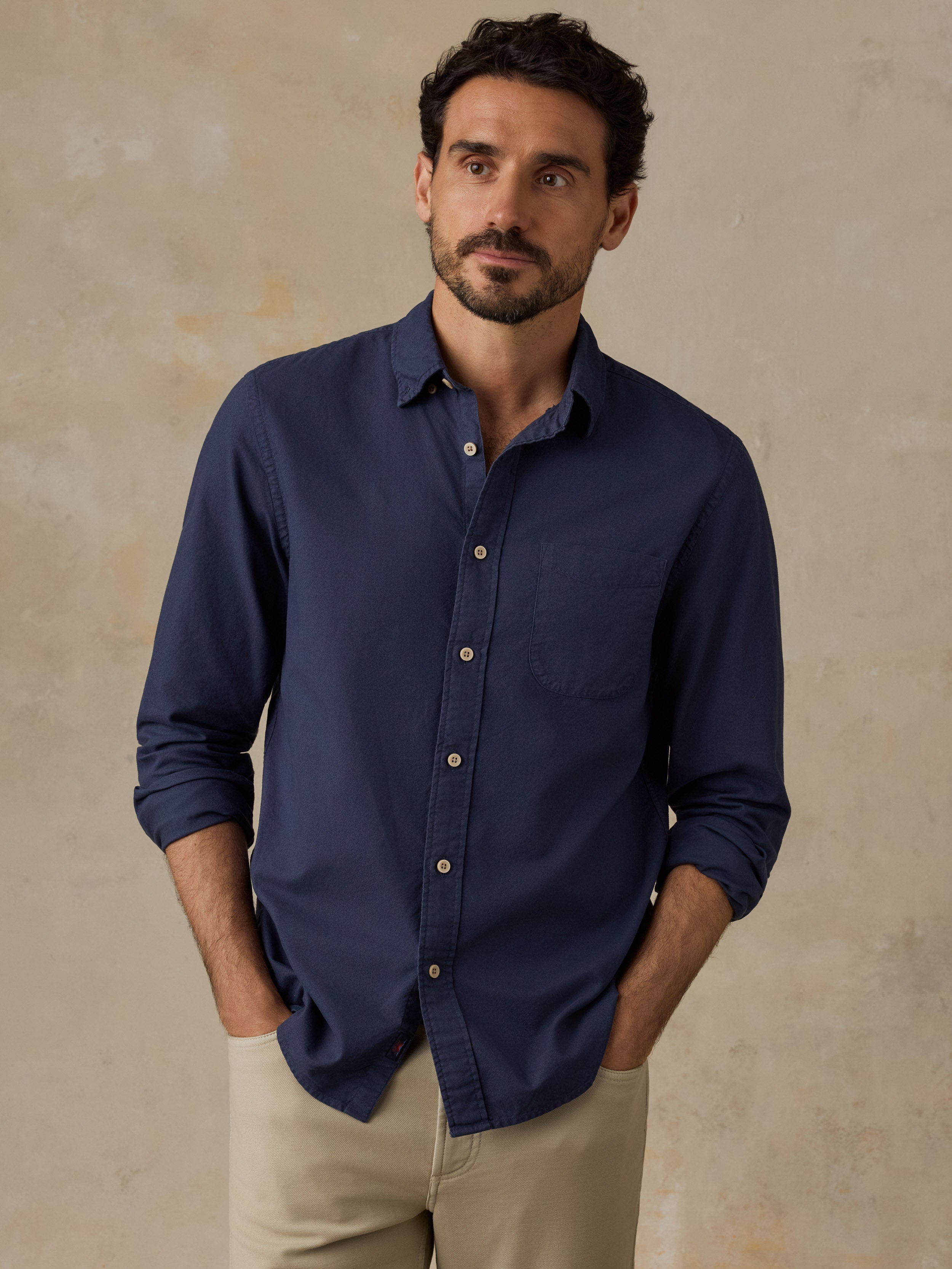 A man wearing a navy blue long-sleeve button-down shirt with rolled-up sleeves and beige trousers, standing against a textured beige wall.