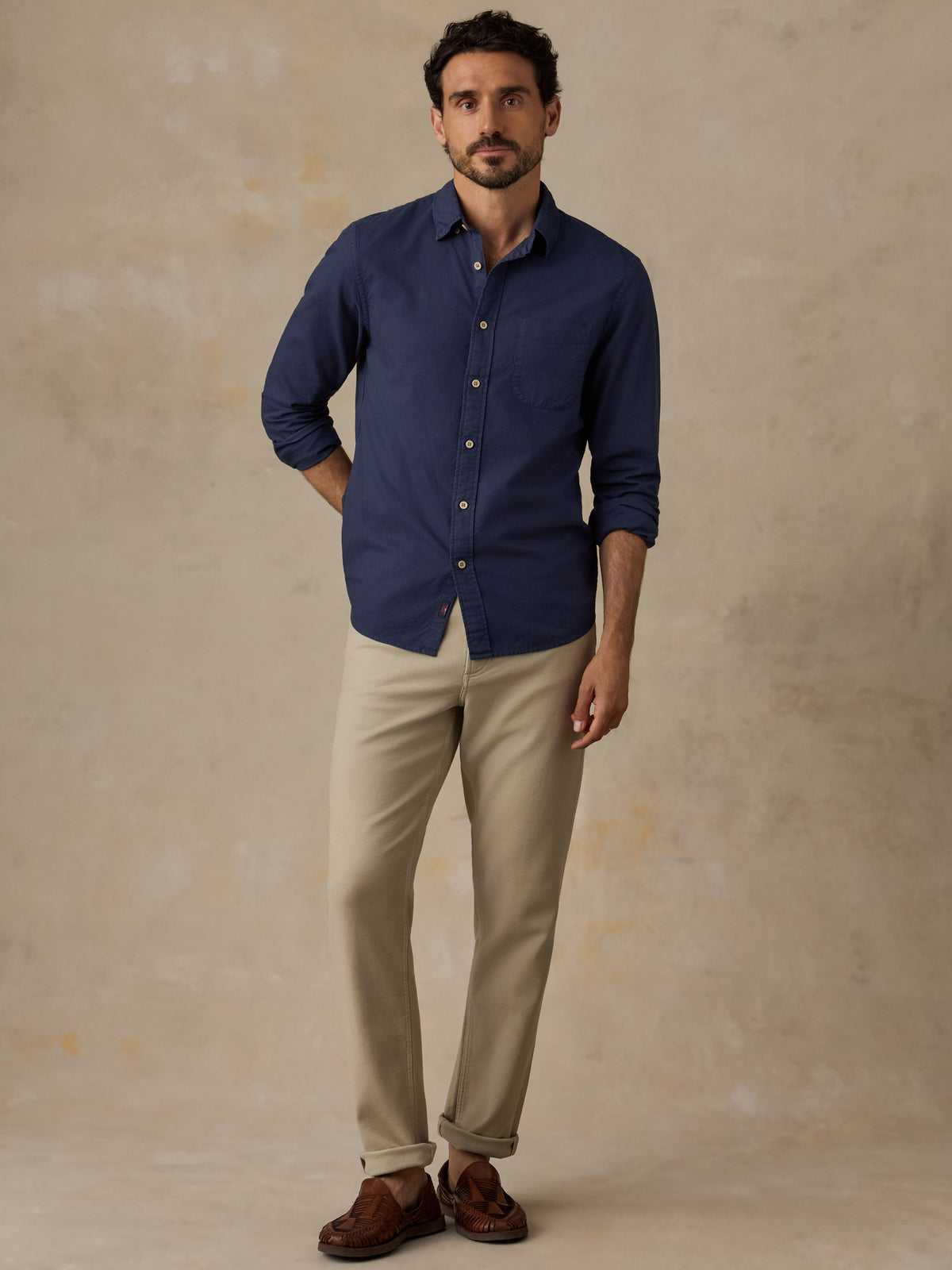 A man wearing a navy blue long-sleeve button-down shirt, beige trousers, and brown leather loafers, standing against a textured beige studio backdrop., image 5 of 9