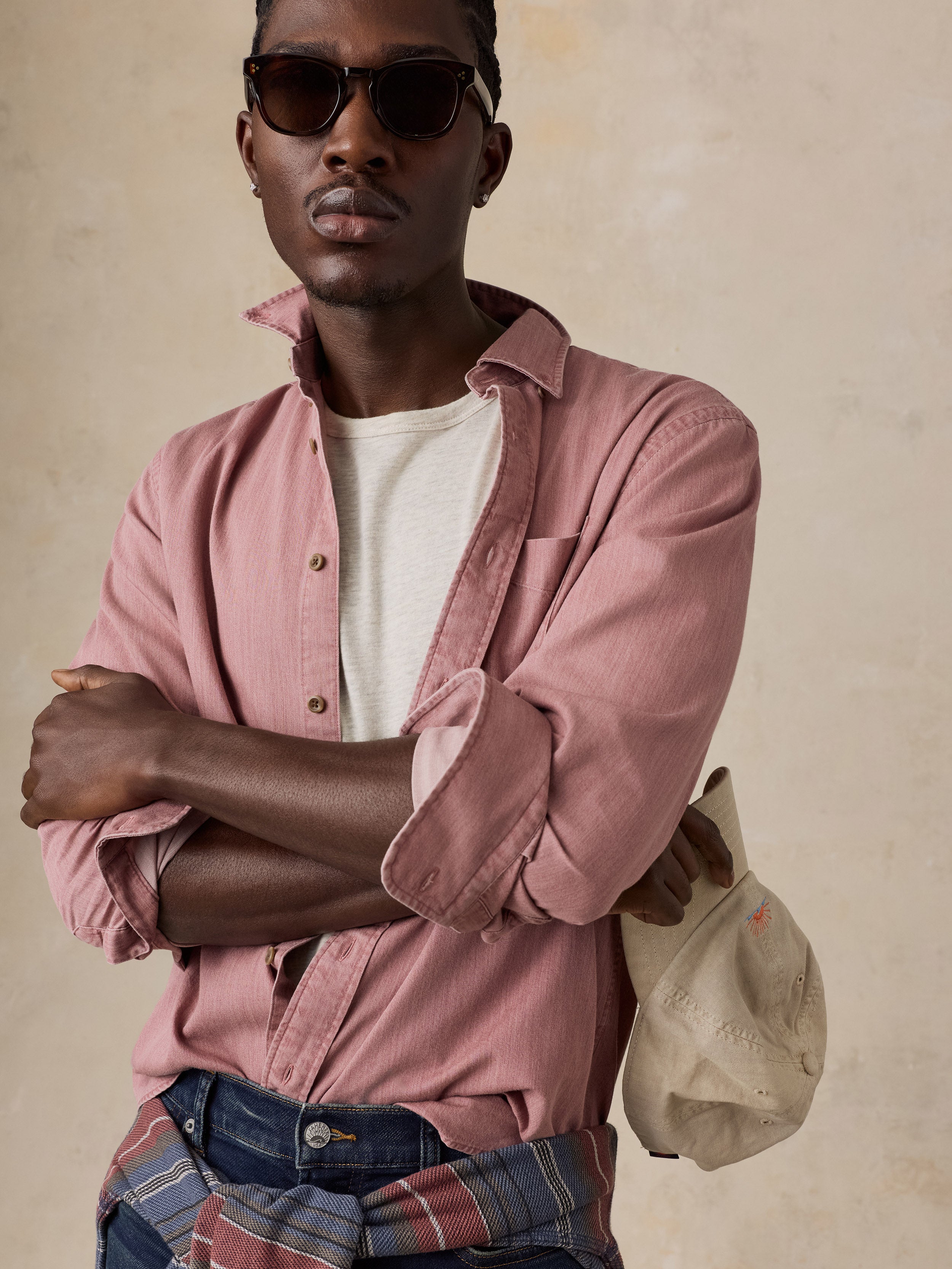 A man wearing a dusty rose pink button-down shirt over a white t-shirt, paired with blue jeans and sunglasses, holding a beige cap against a textured beige background.