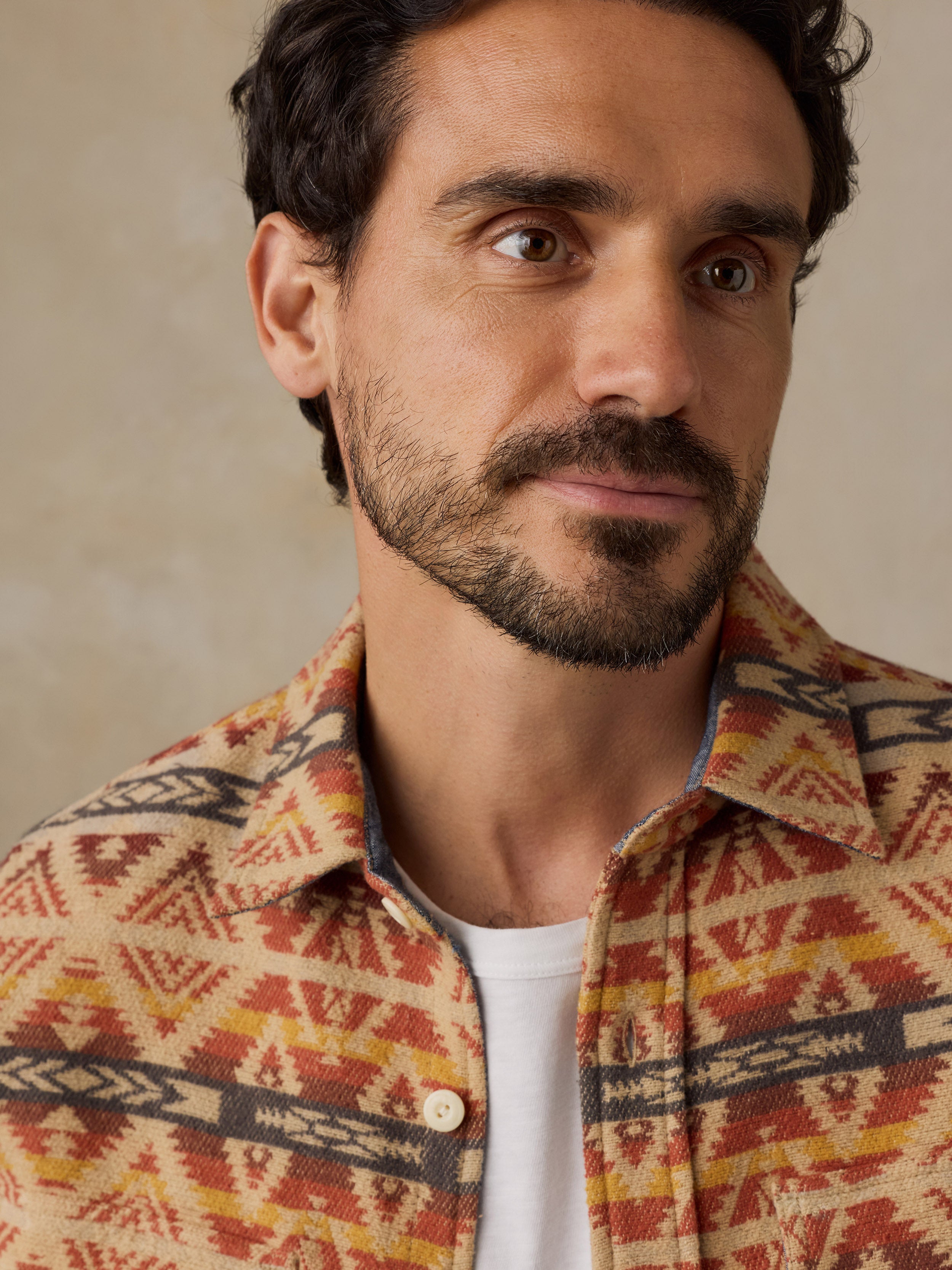 A close-up portrait of a man wearing a collared button-up shirt with a Southwestern geometric pattern in orange, yellow, and dark brown over a white t-shirt, set against a neutral beige background.