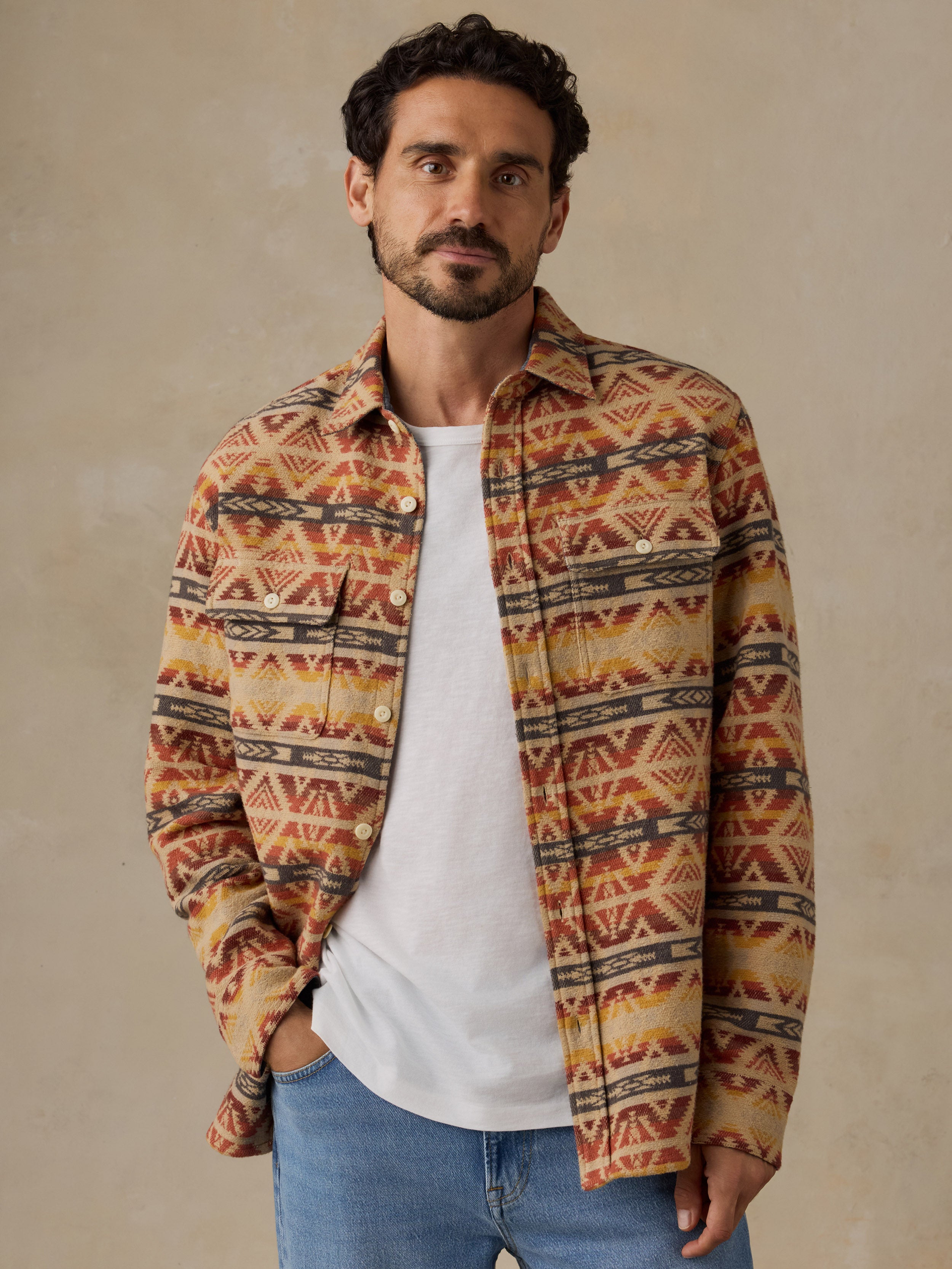 A man wearing a long-sleeve button-up shirt with a Southwestern geometric pattern in orange, brown, and beige, layered over a white t-shirt and blue jeans, standing against a neutral textured beige wall.