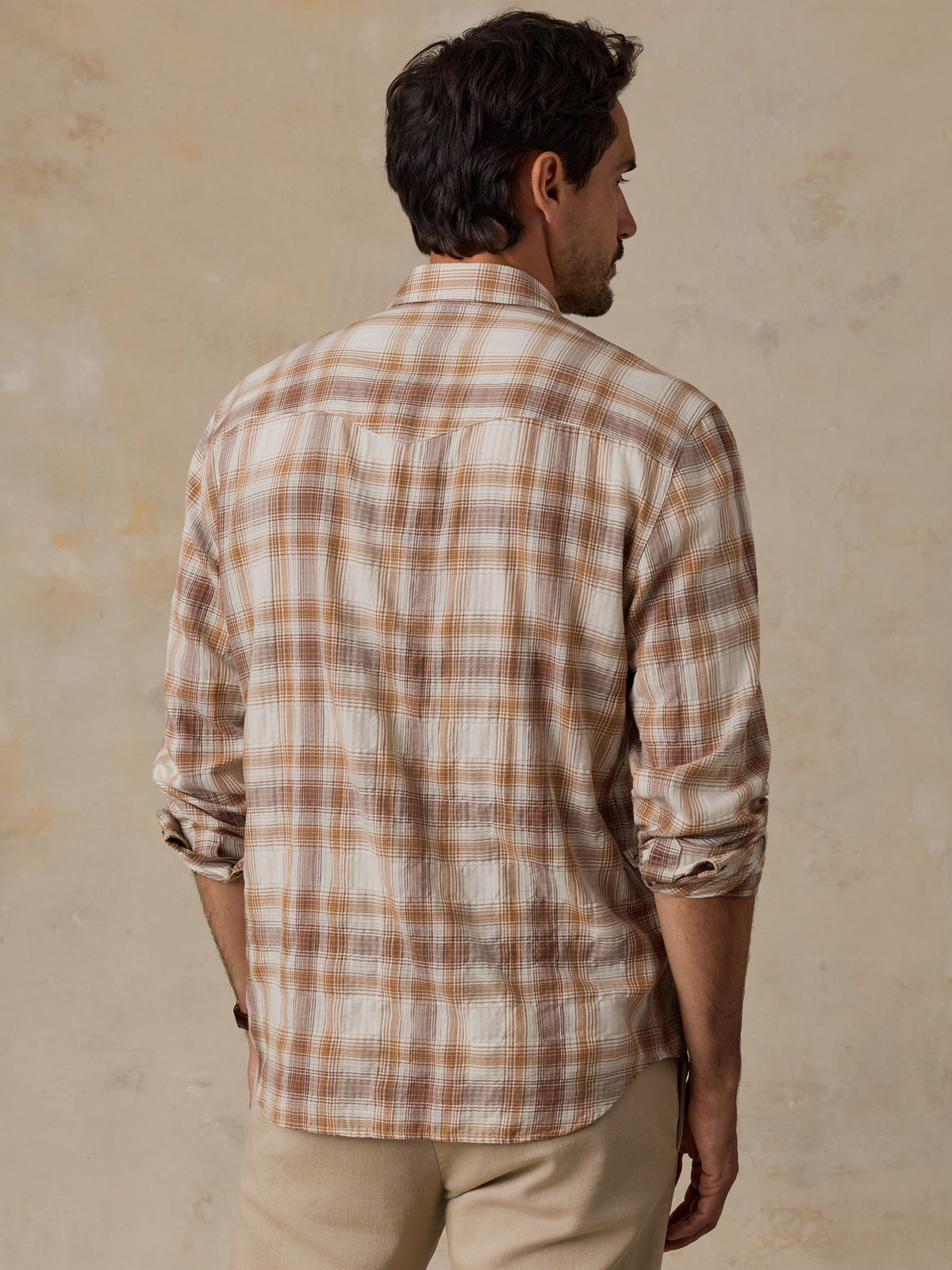 A man viewed from behind wearing a long-sleeve plaid button-up shirt in beige, brown, and white, paired with khaki pants, set against a textured beige wall.