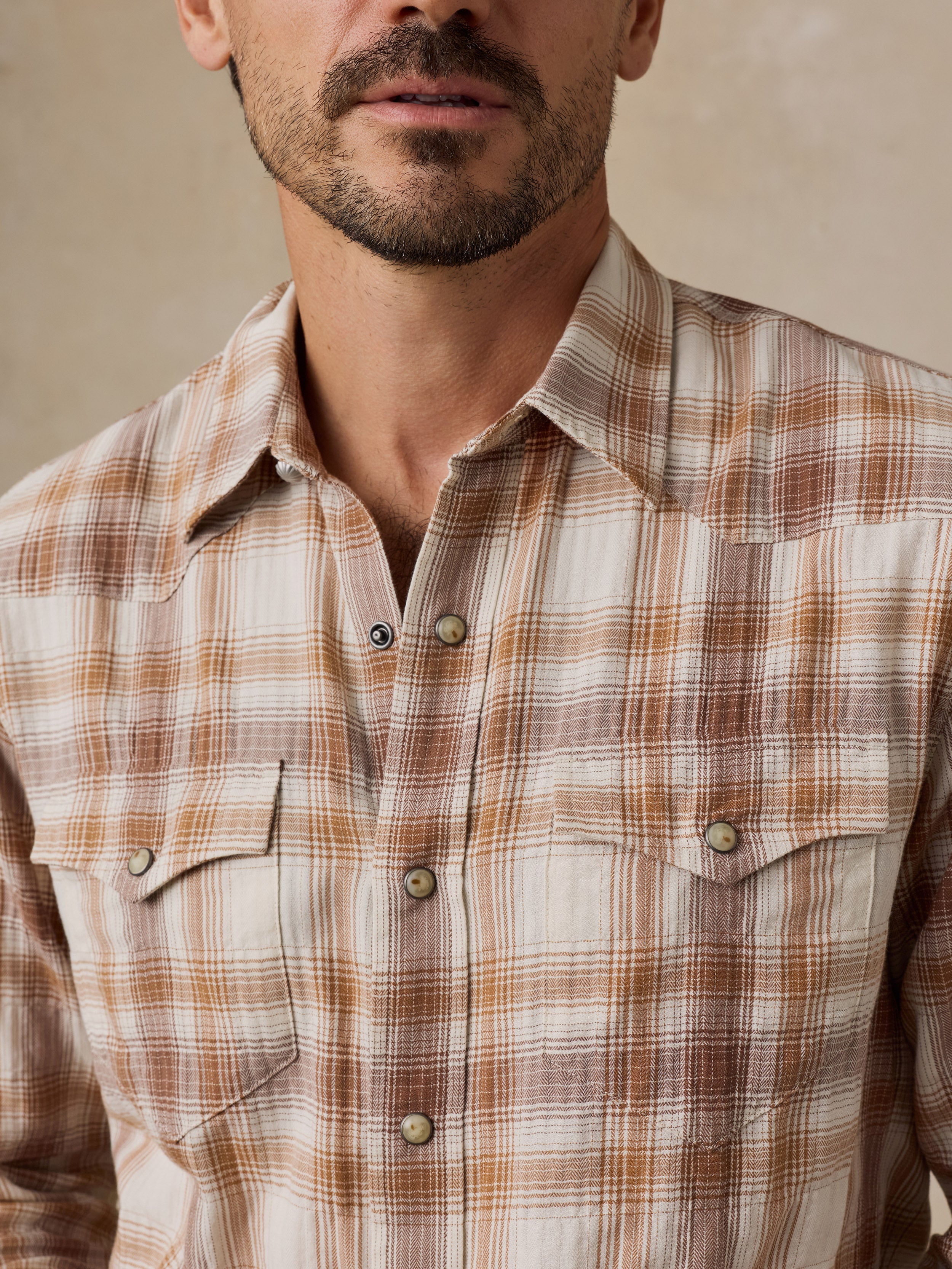 A close-up of a man wearing a brown and beige plaid western-style button-down shirt with two chest pockets.