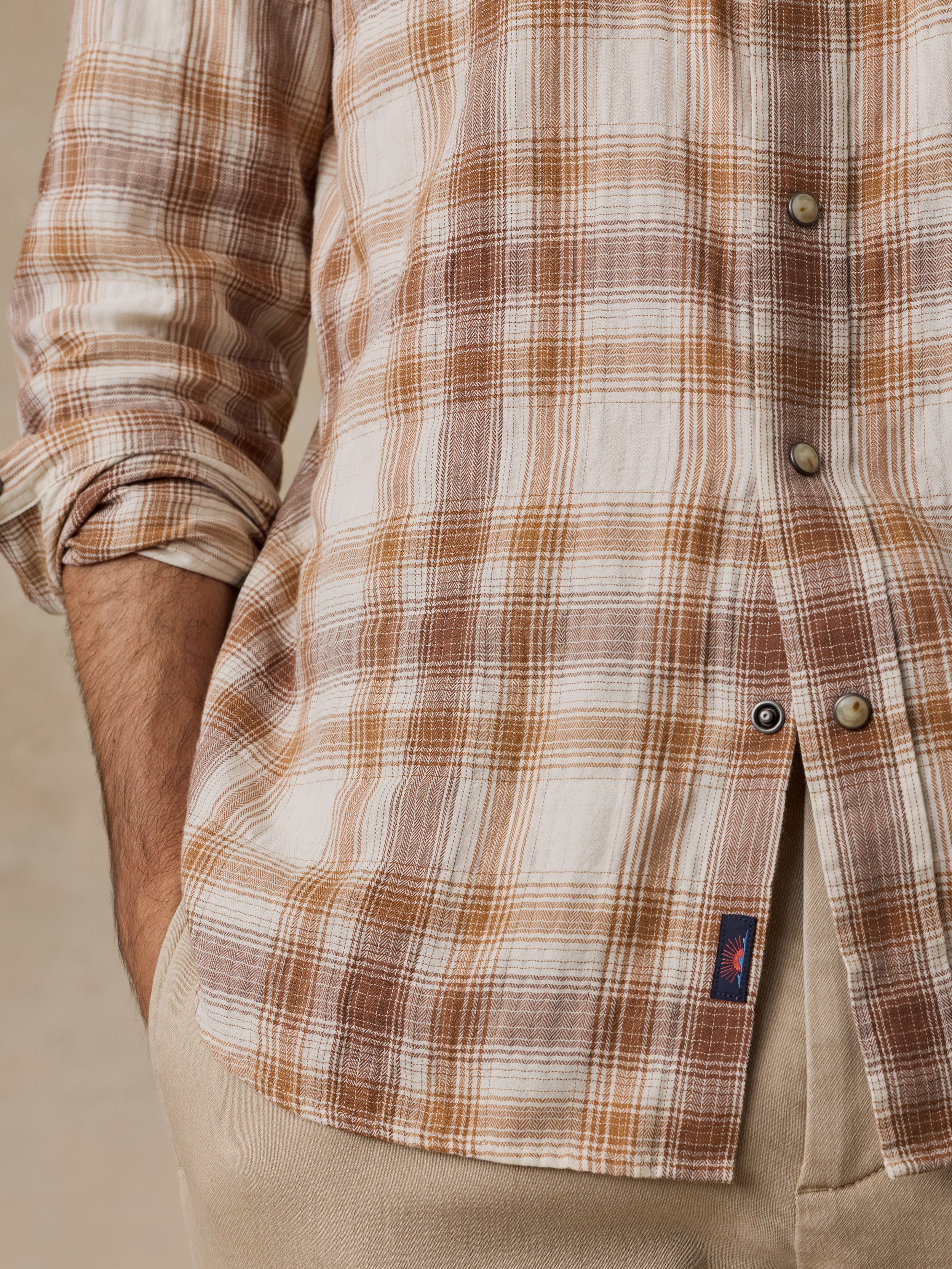 Close-up of a man wearing a long-sleeve plaid button-up shirt in brown, beige, and white tones, paired with khaki pants.
