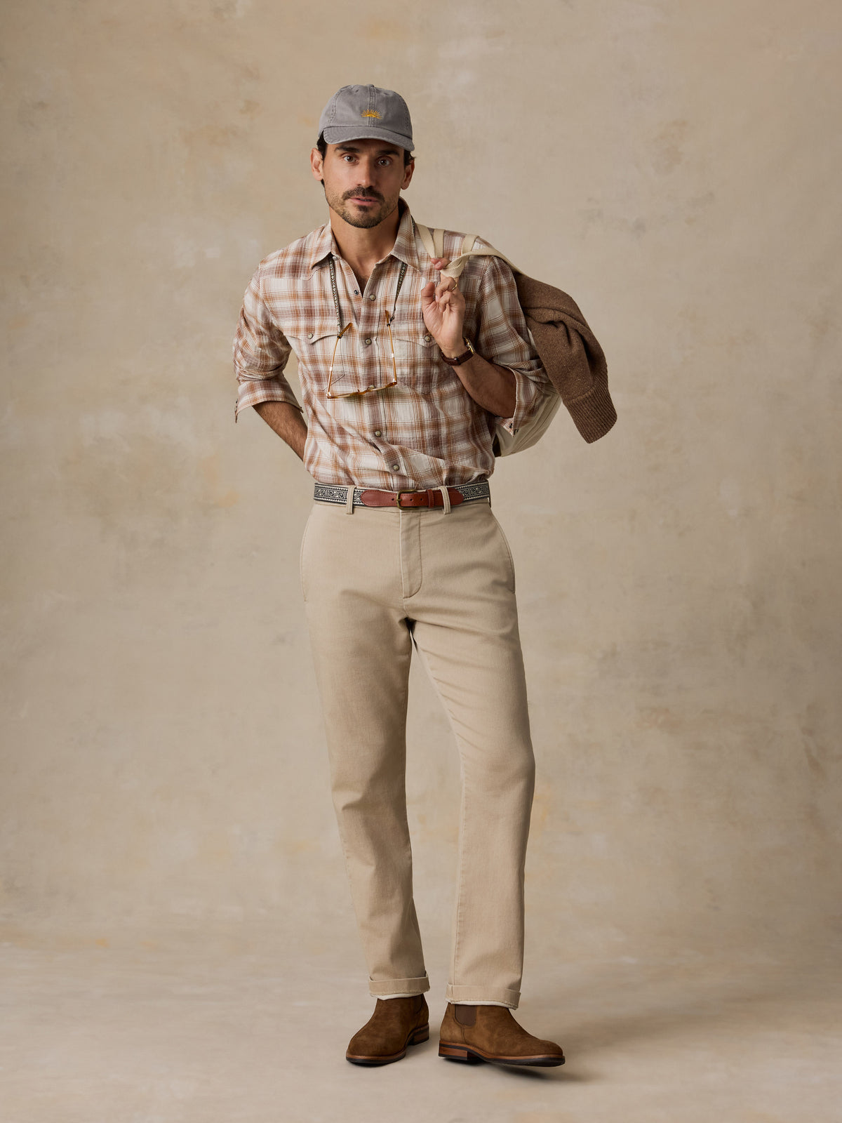 A man stands against a textured beige studio backdrop wearing a brown and beige plaid button-down shirt, khaki pants, brown boots, and a gray cap, with a brown jacket slung over his shoulder.