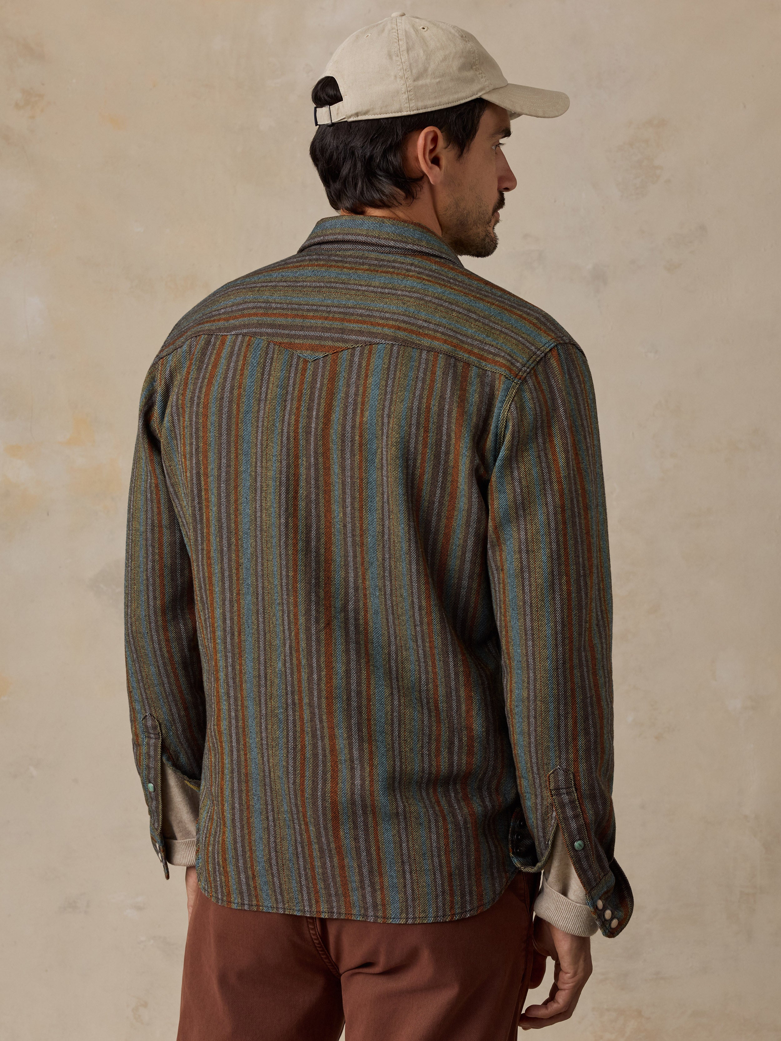 A man wearing a striped button-down shirt with blue, green, and brown vertical stripes, paired with brown pants and a beige baseball cap.