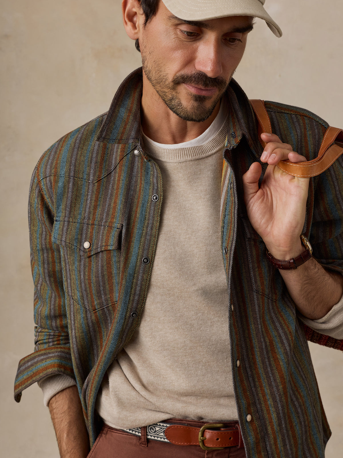 A man wearing a striped button-down shirt with blue, green, and brown vertical stripes, paired with brown pants and a beige baseball cap