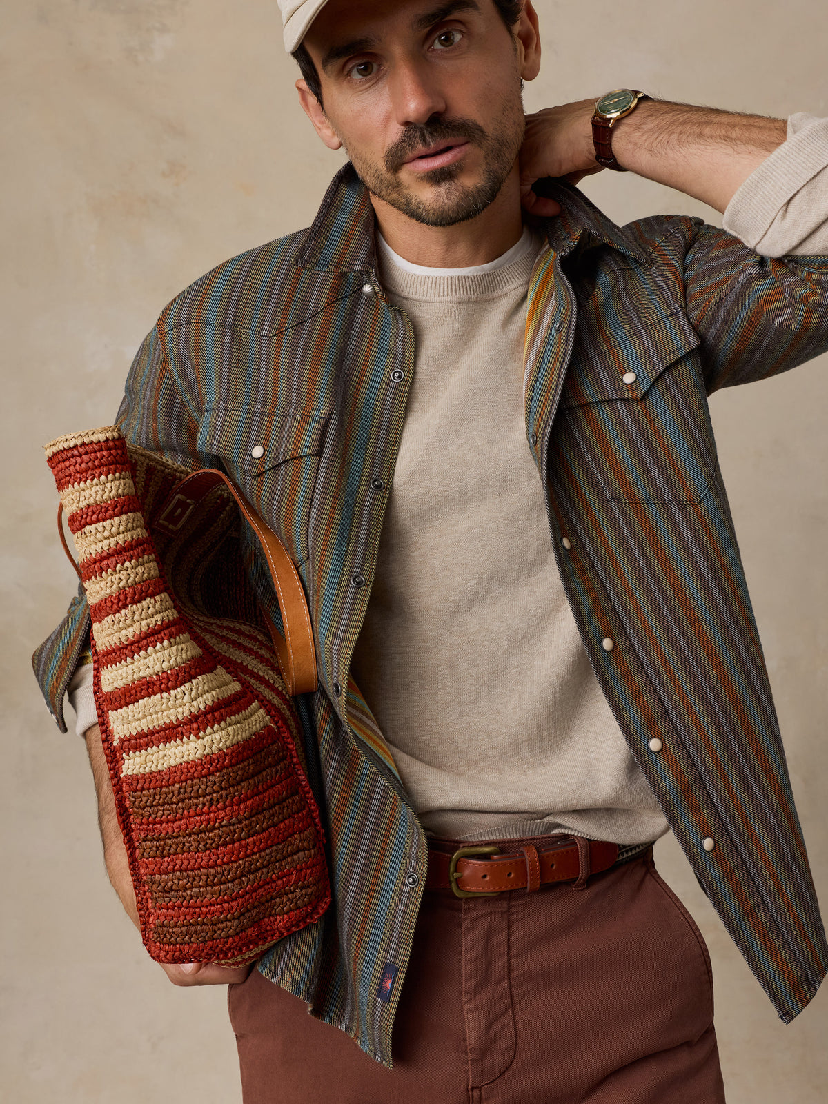 A man wearing a striped button-down shirt with blue, green, and brown vertical stripes, paired with brown pants and a beige baseball cap