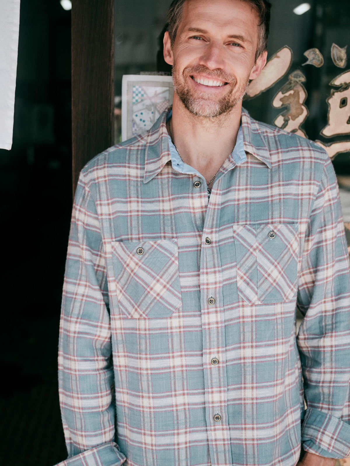 A man wearing a long-sleeve plaid button-up shirt in muted blue, beige, and red tones, image 1 of 7