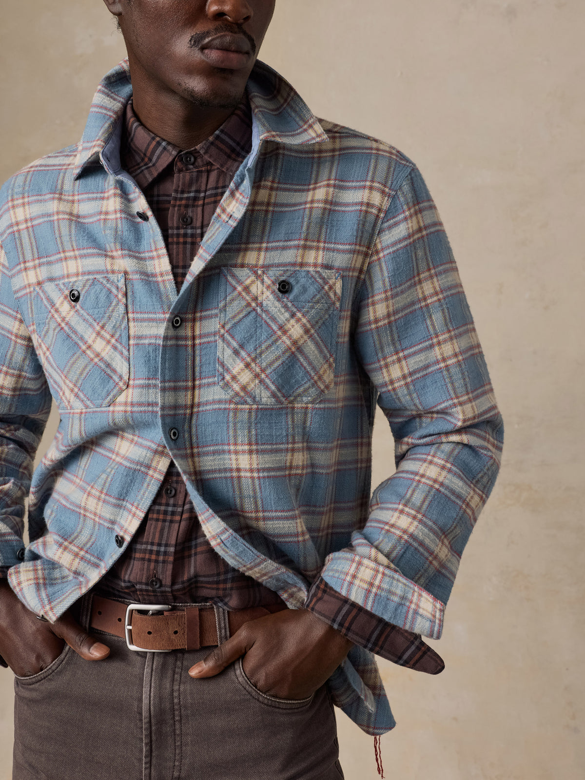 A man wearing a light blue plaid shirt over a plaid button up with brown pants, image 4 of 7