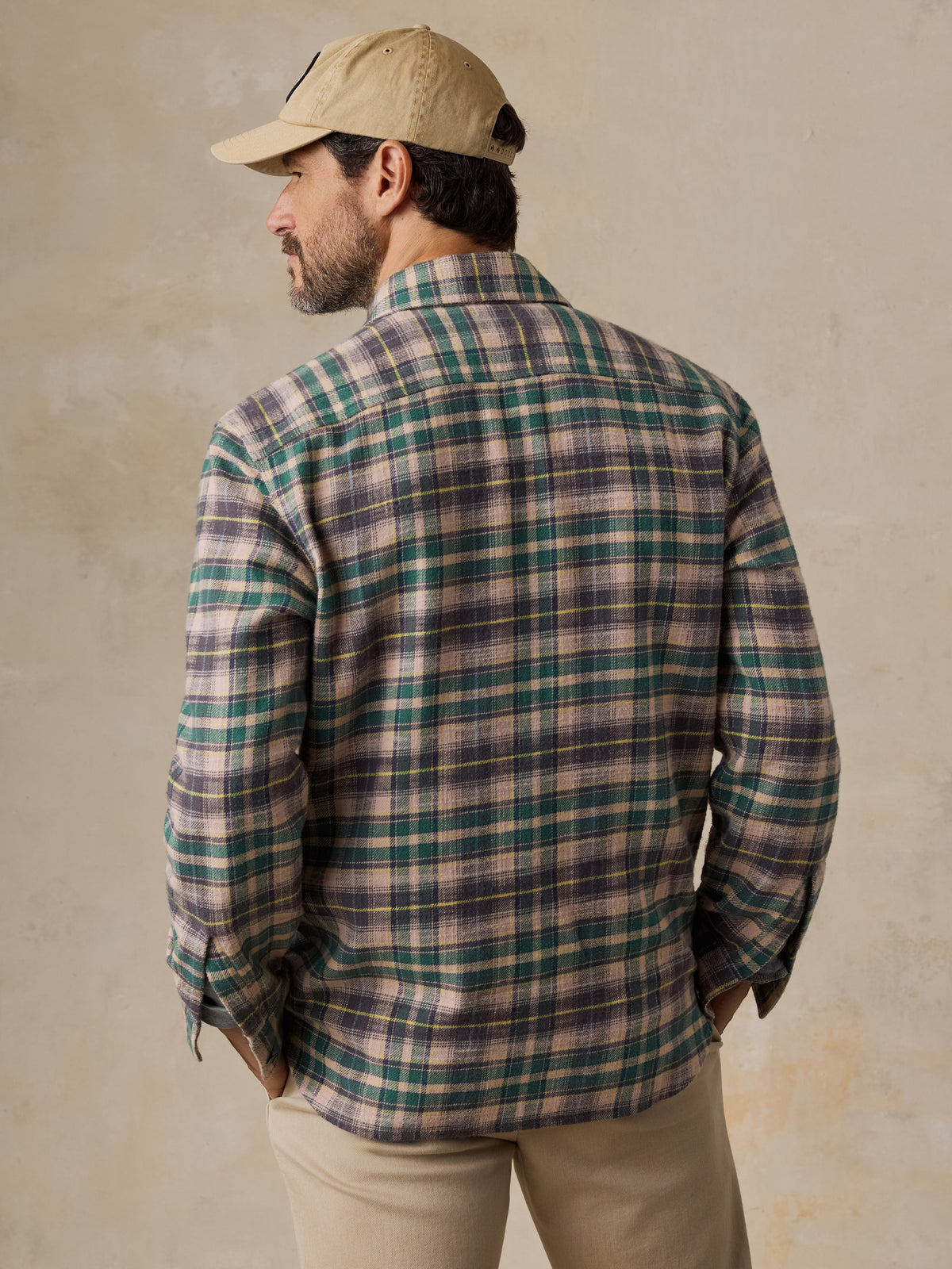 A man viewed from behind wearing a green flannel with beige pants and a hat, image 2 of 5