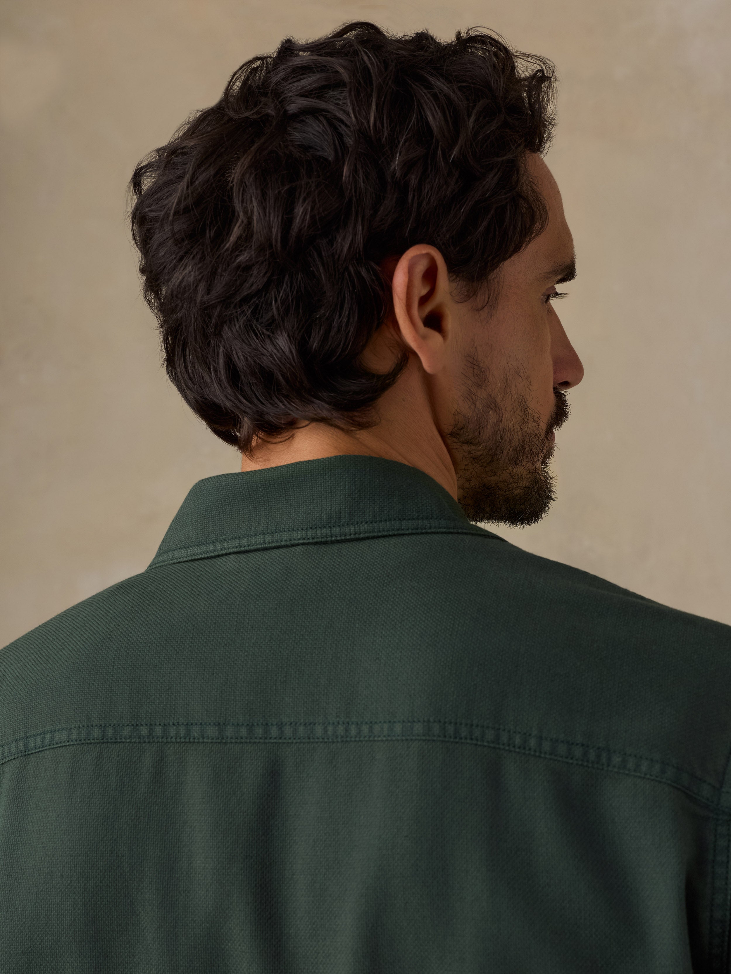 A man shown from the back and side profile wearing a dark forest green button-down shirt against a textured beige background.