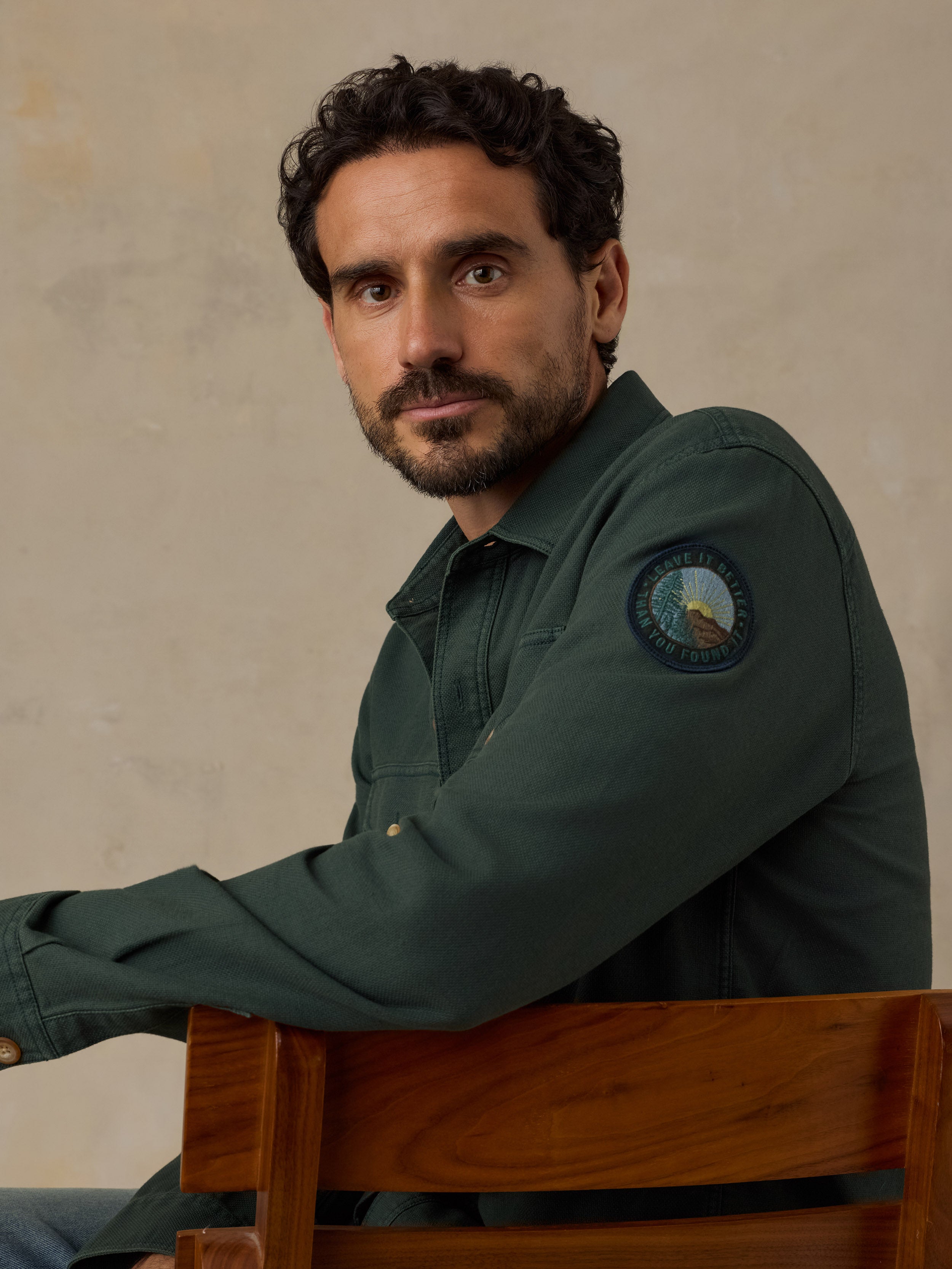 A man with dark curly hair and a beard wearing a dark green button-up shirt with a circular 'Leave It Better Than You Found It' patch on the sleeve, sitting on a wooden chair against a textured beige wall.