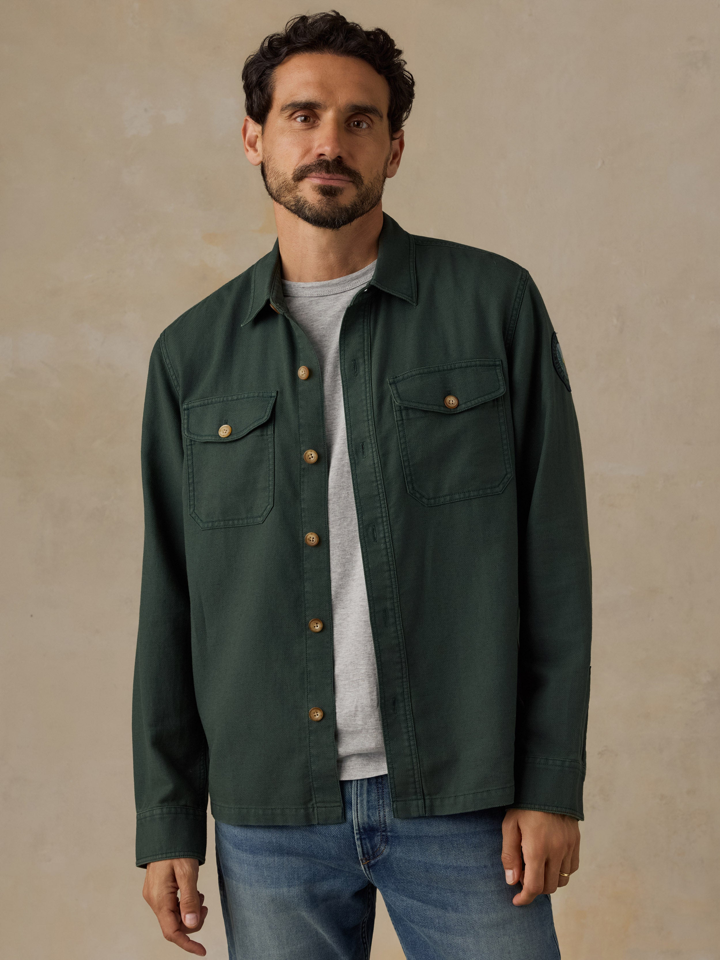 A man wearing a dark green button-up overshirt with two chest pockets and brown buttons, layered over a light gray t-shirt and blue jeans, standing against a textured beige background.