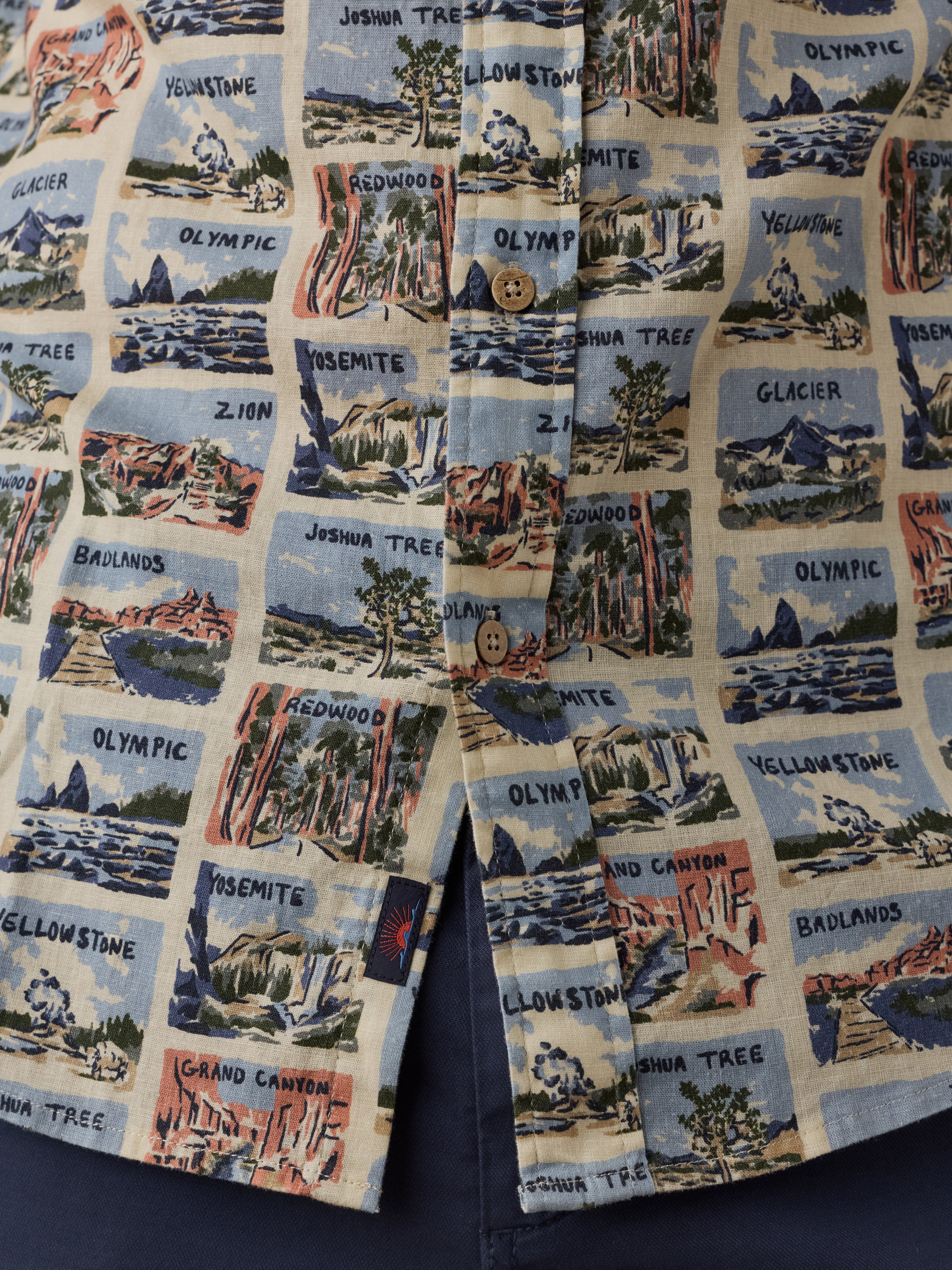 A close-up of a button-front garment, likely a shirt or jacket, featuring an all-over printed pattern of illustrated canyon and mountain scenes with labels such as Yellowstone, Grand Canyon, Zion, and Olympic. The print is set on a light cream background with blue, red, green, and brown scenic illustrations.