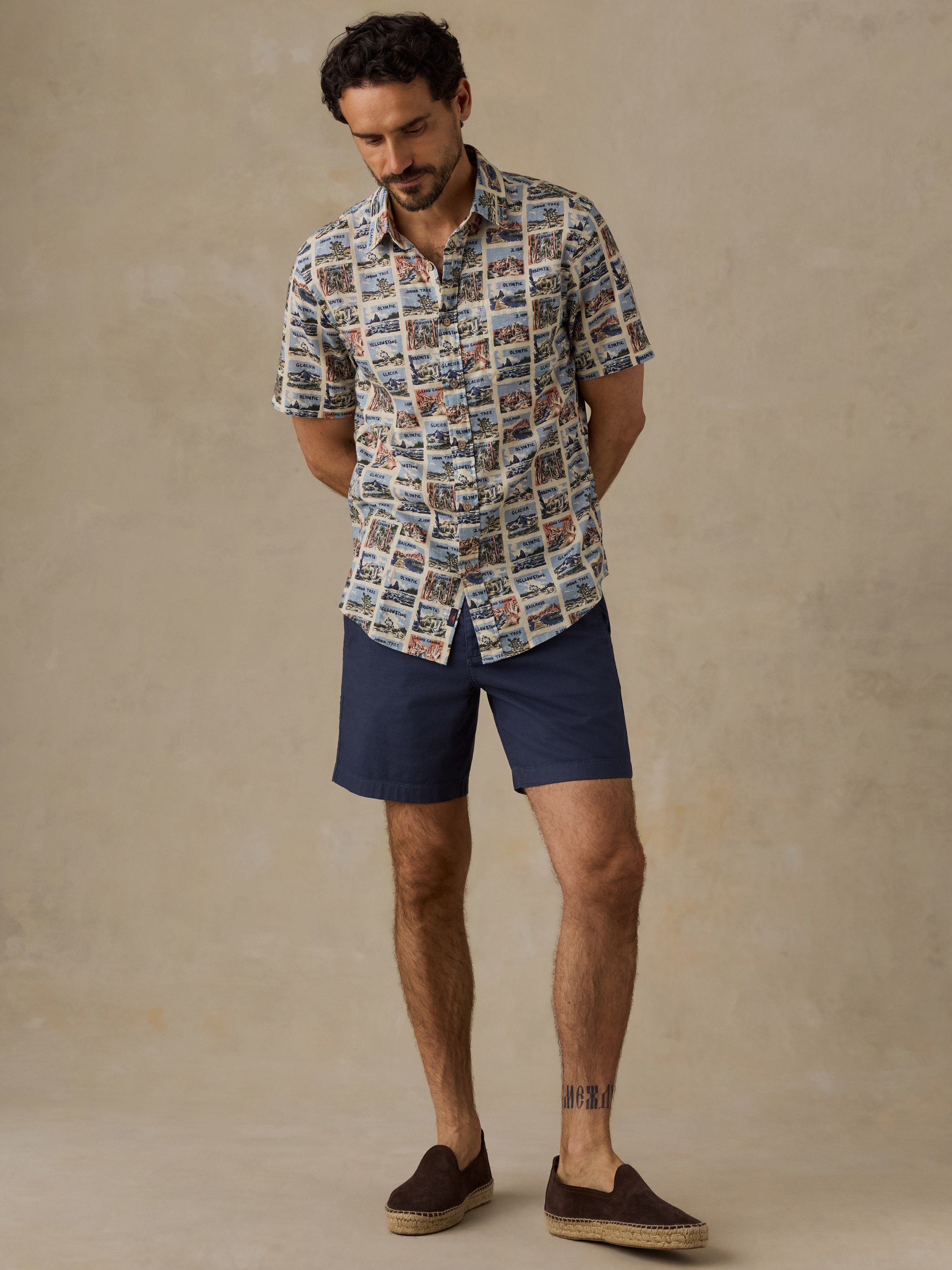 A man wearing a short-sleeve button-up shirt with a detailed scenic print, navy blue shorts, and brown espadrille loafers, standing against a textured beige studio backdrop.