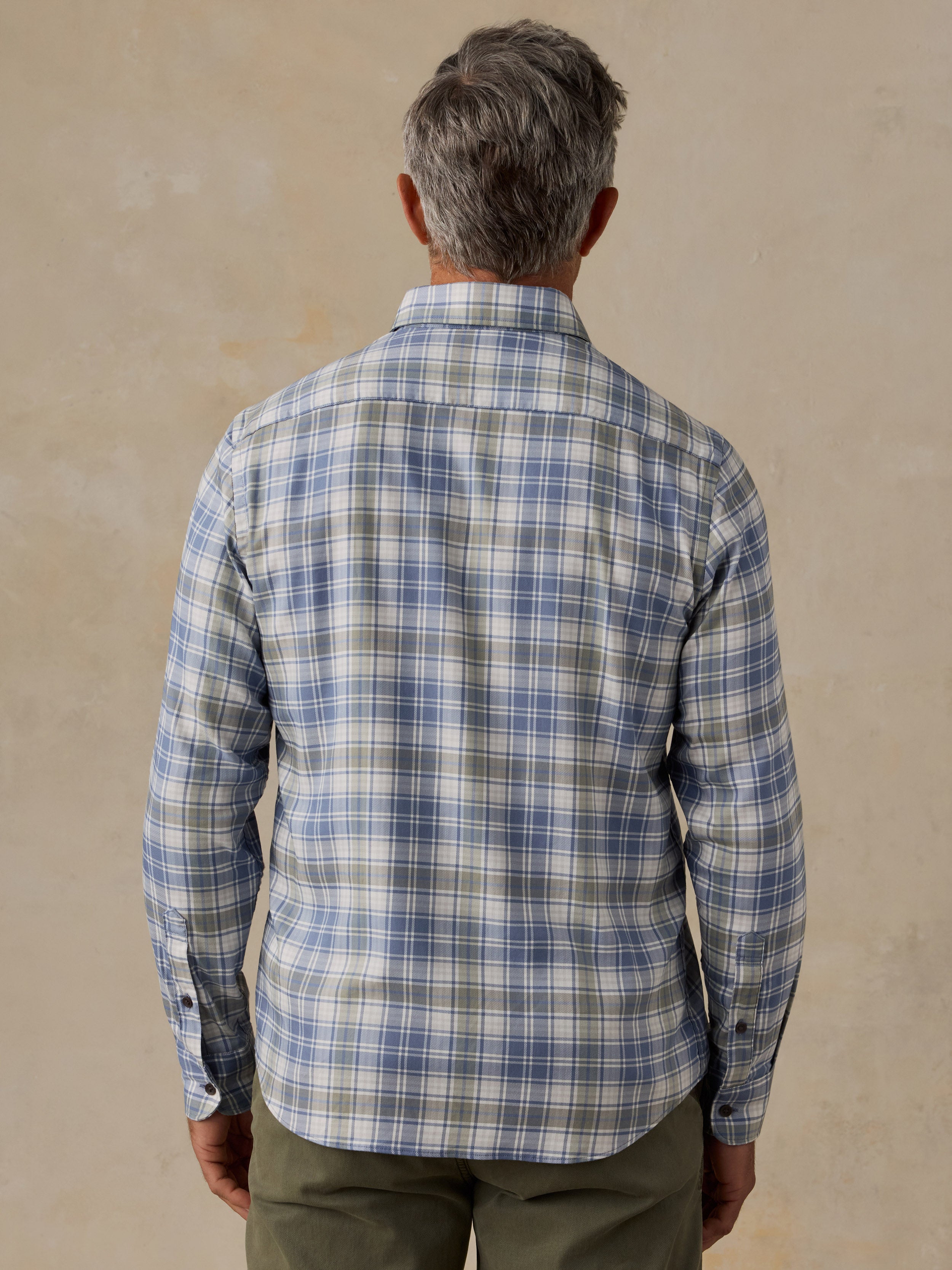 A man viewed from behind wearing a long-sleeve plaid button-down shirt in blue, gray, and white, paired with olive green pants, set against a textured beige wall.