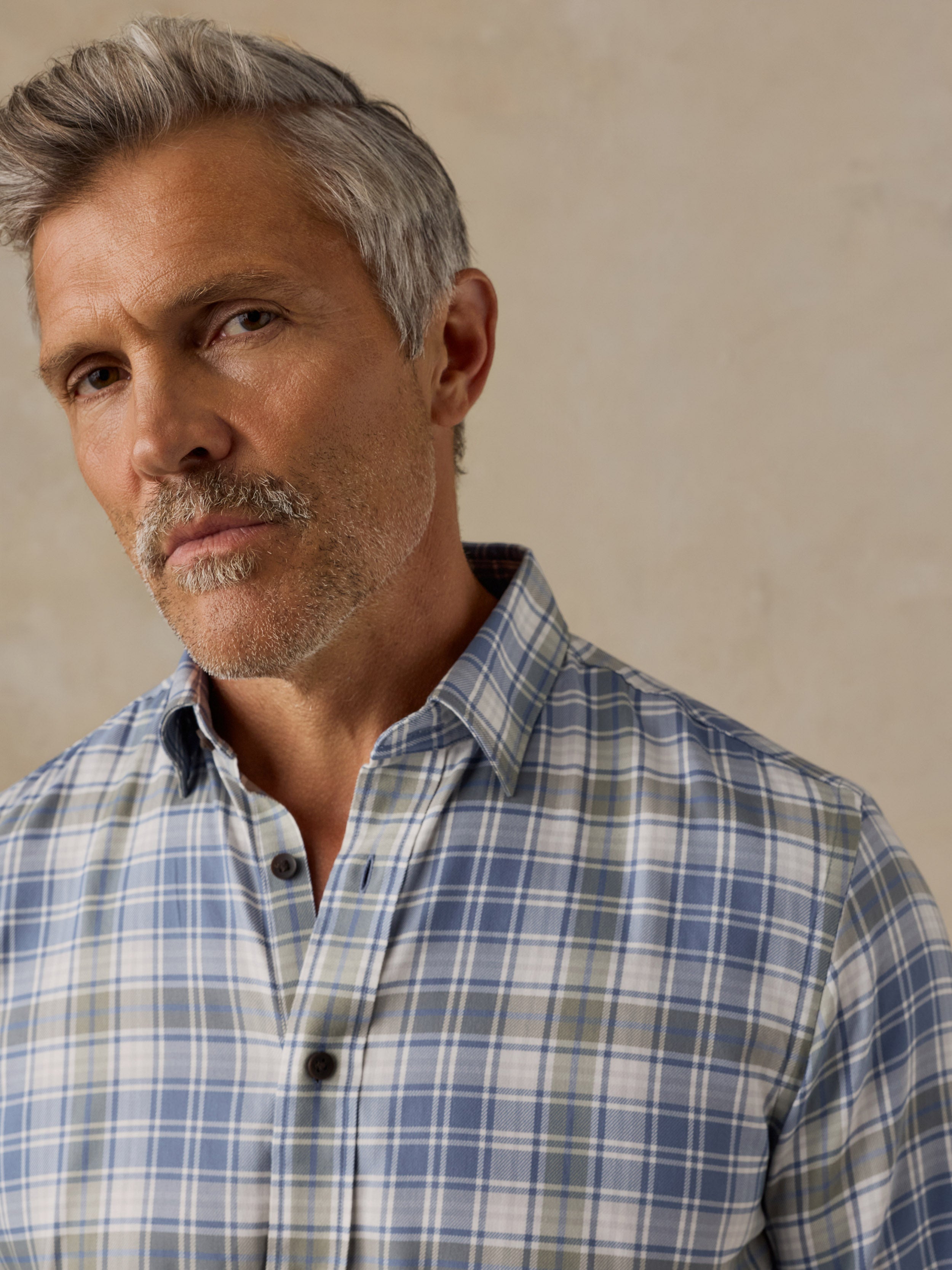 A close-up portrait of a man with gray hair and a mustache wearing a blue, white, and light green plaid button-down shirt against a textured beige background.