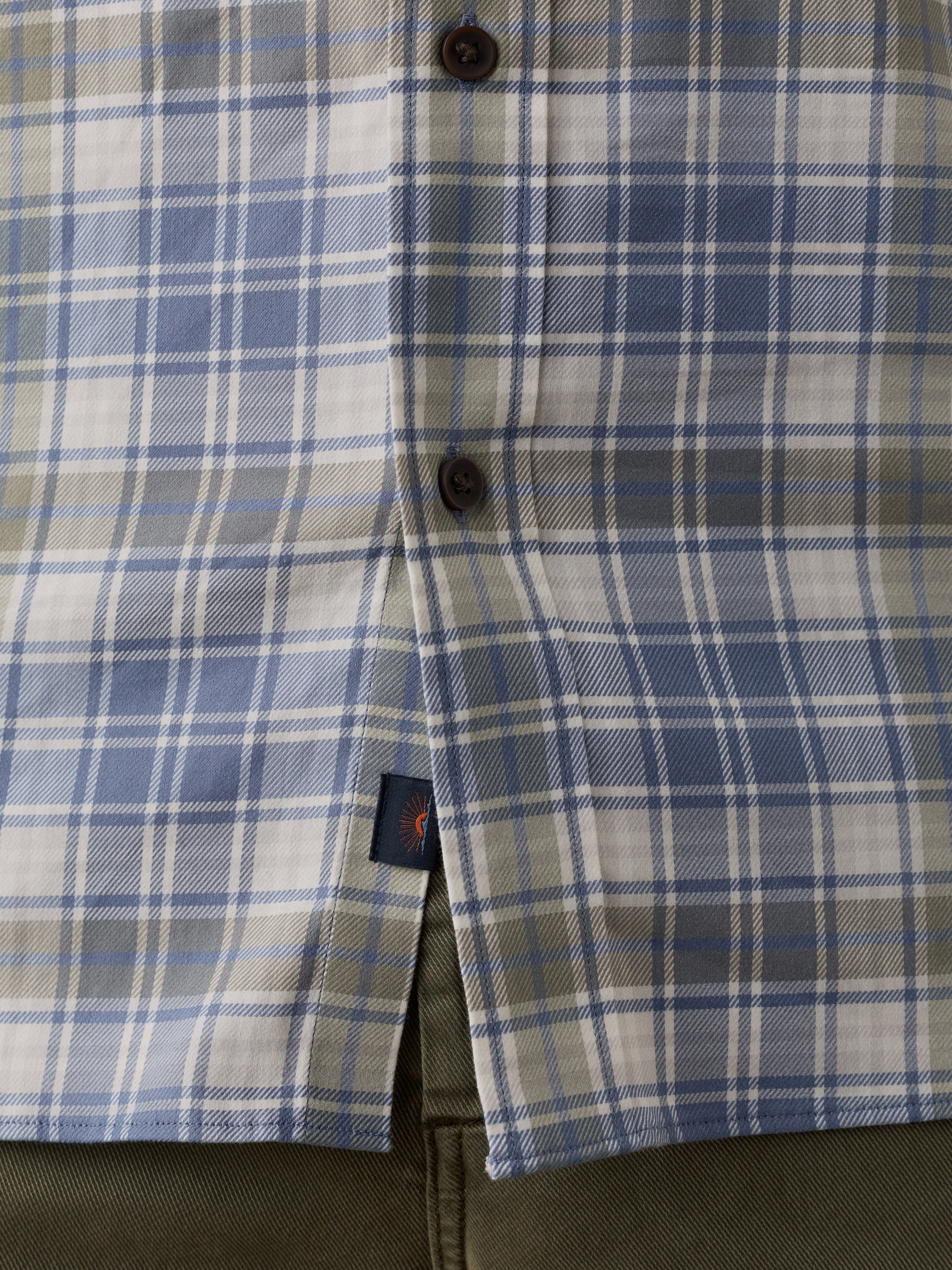 Close-up of a plaid button-up shirt in blue, white, and gray, with dark buttons and a small Faherty brand tag near the hem.