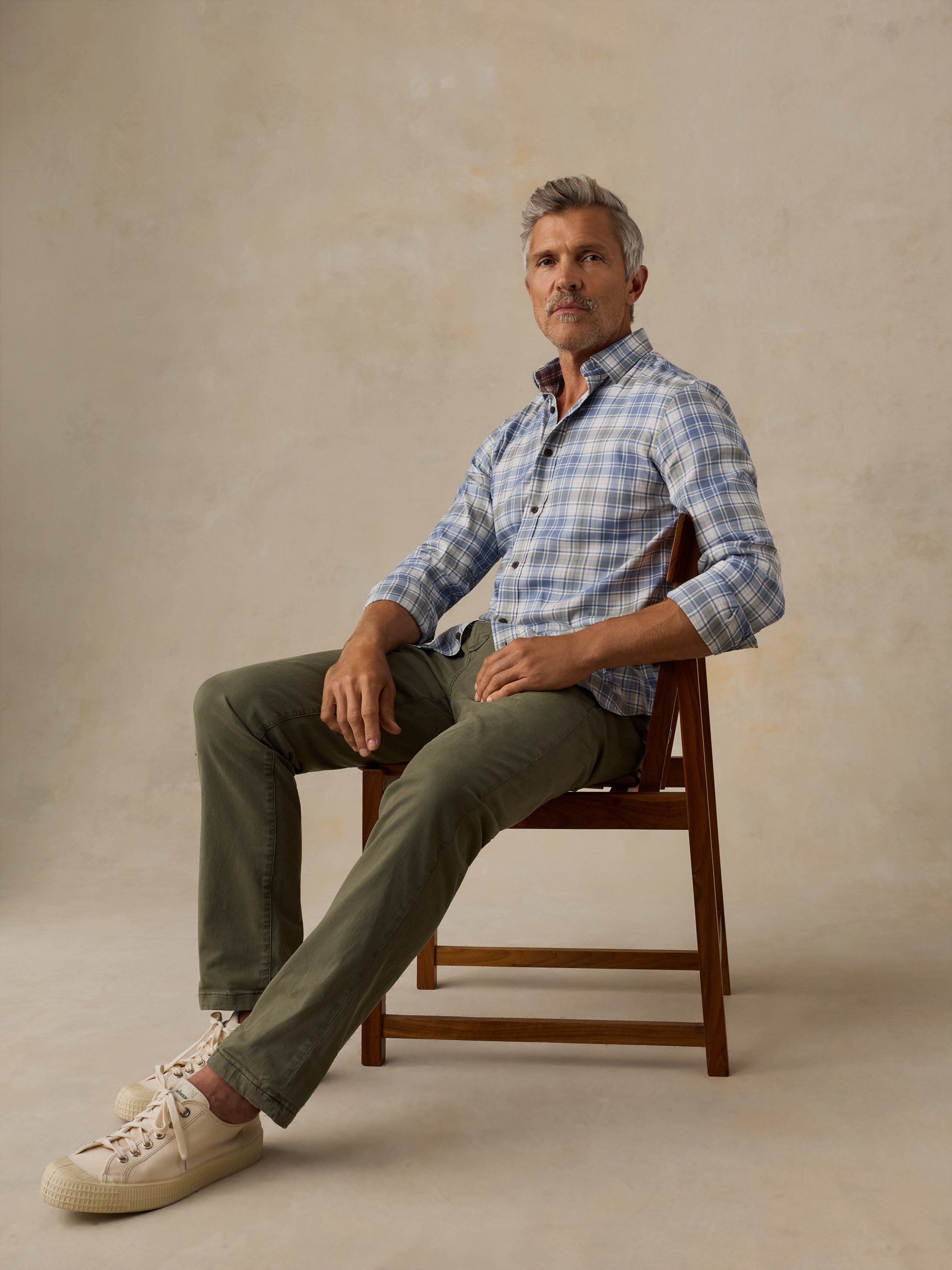 A man sits on a wooden chair wearing a light blue and white plaid button-down shirt, olive green pants, and cream-colored sneakers against a textured beige studio backdrop.