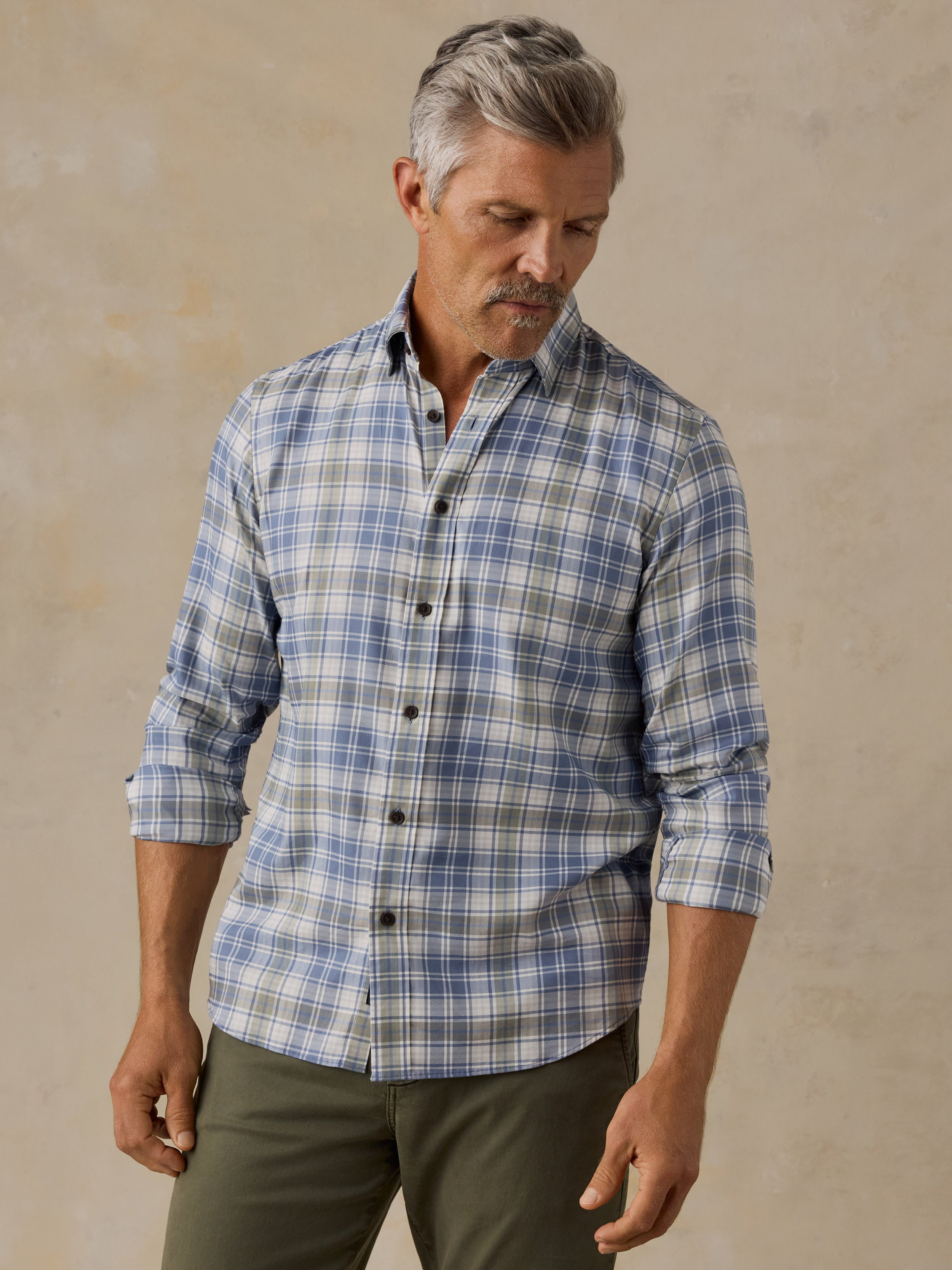 A man wearing a blue and white plaid button-down shirt with olive green pants, standing against a textured beige background.