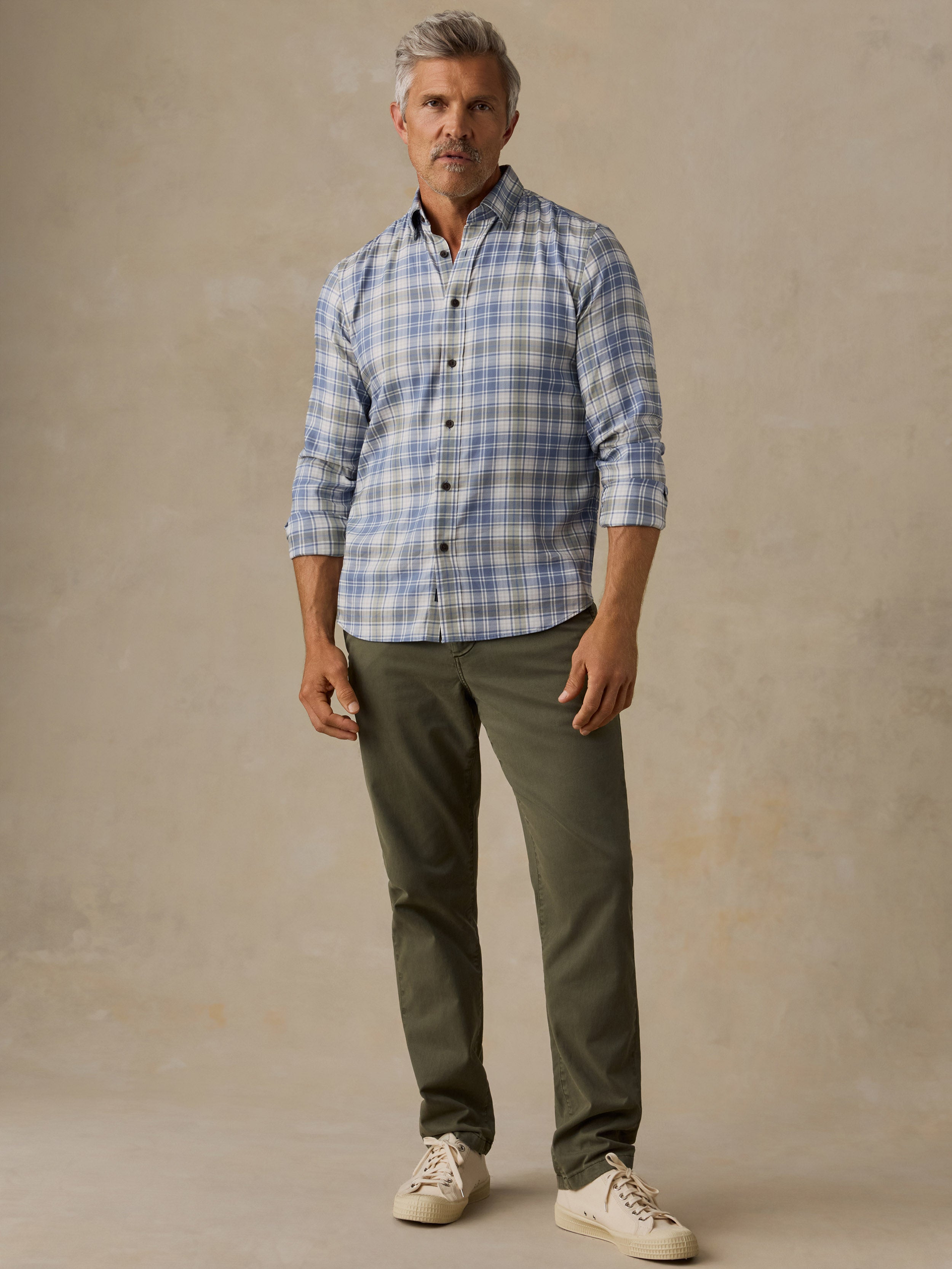 A man wearing a light blue and white plaid button-down shirt, olive green trousers, and off-white sneakers, standing against a textured beige studio backdrop.