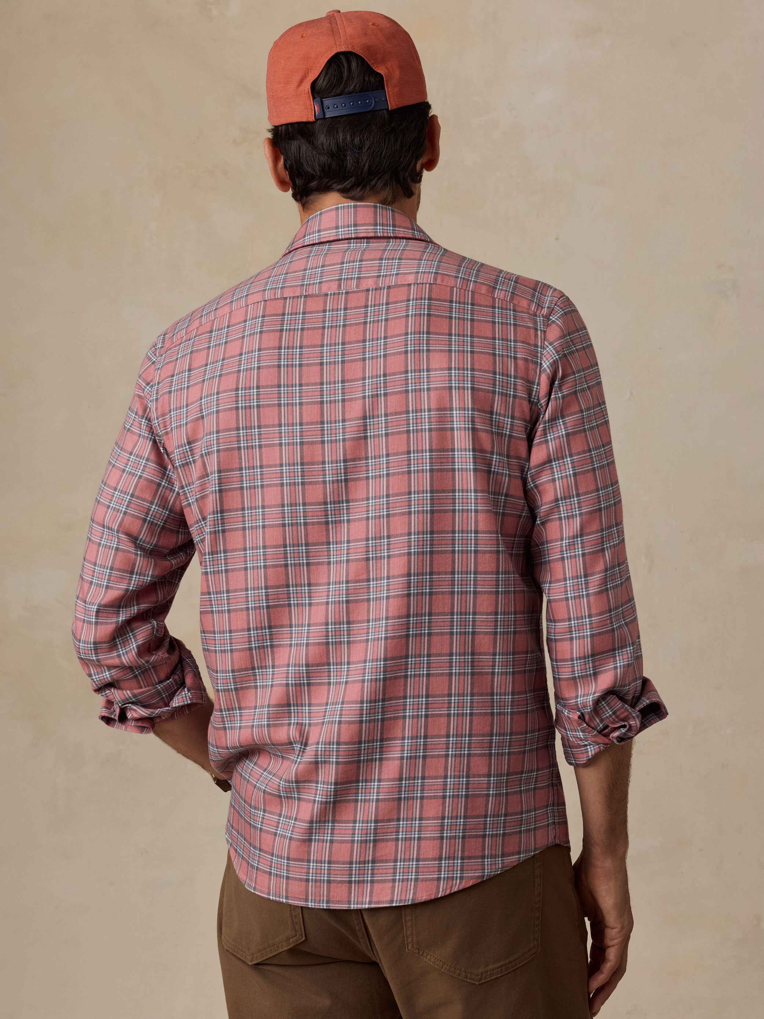 A man wearing a pink and blue plaid button-down shirt, brown pants, and an orange cap.