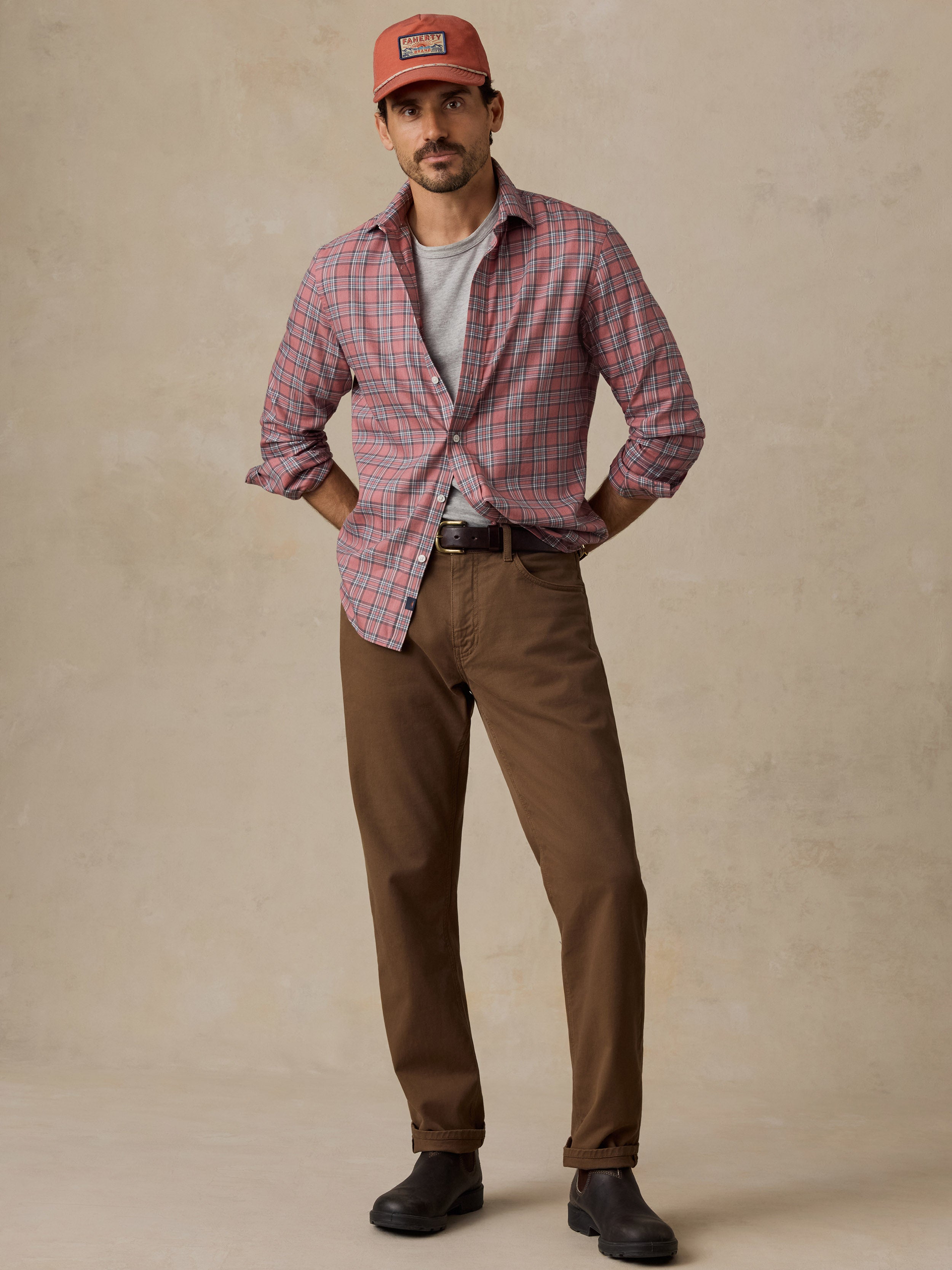 A man wearing a casual outfit consisting of a red and blue plaid button-down shirt, a light gray t-shirt, brown trousers, and an orange cap.