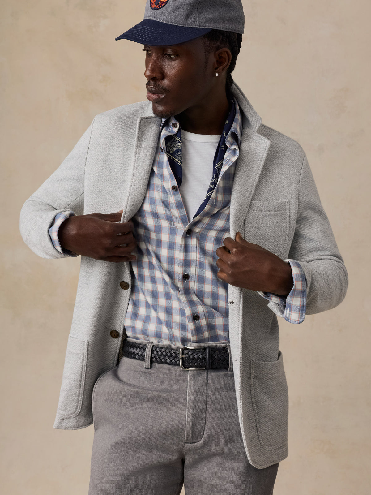 A man wearing a light gray blazer over a blue and white checkered shirt, a white undershirt, gray trousers, and a gray cap.