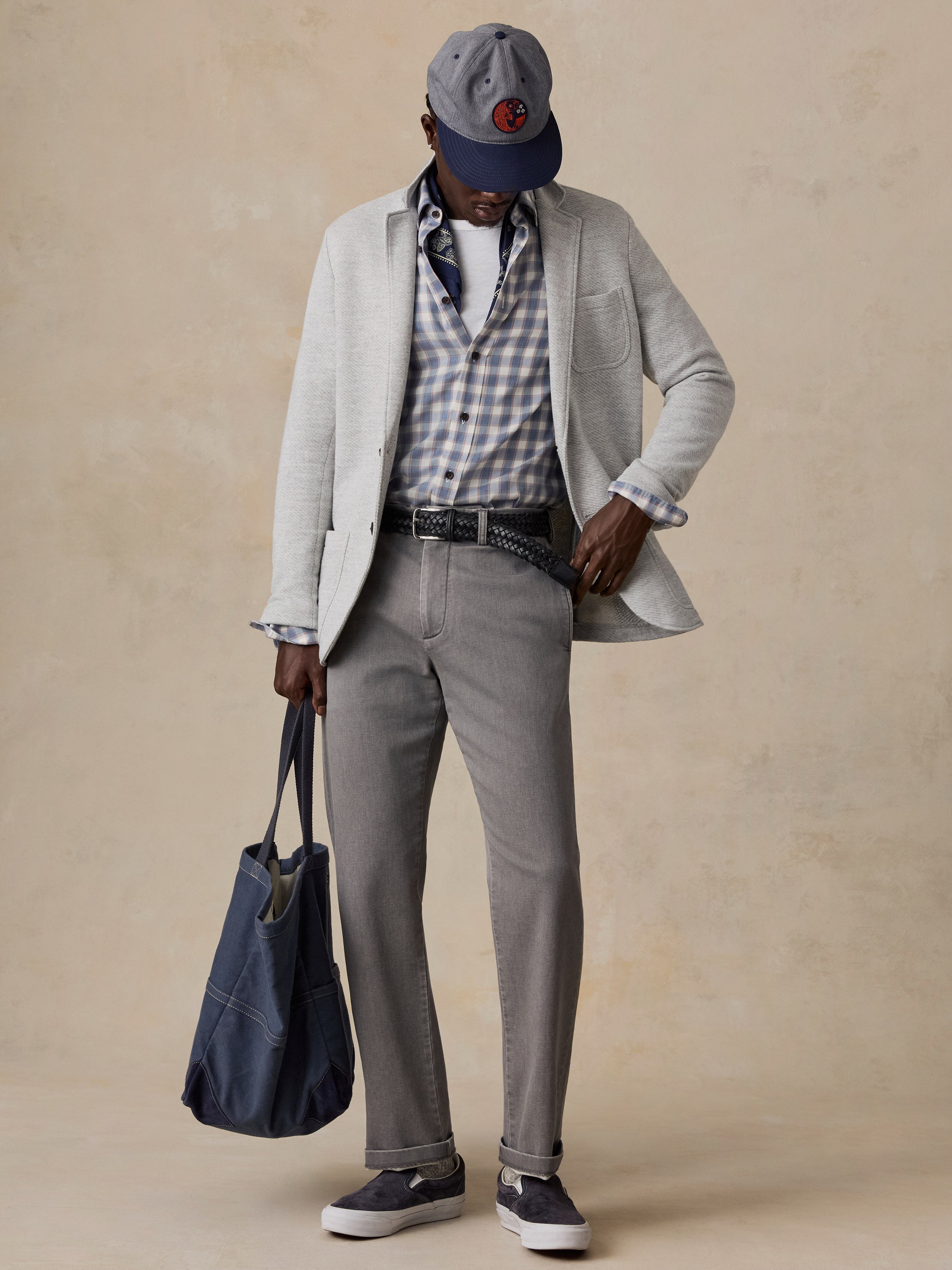 A man wearing a light gray blazer, gray trousers, a blue and white check shirt, and a navy baseball cap, holding a navy tote bag.