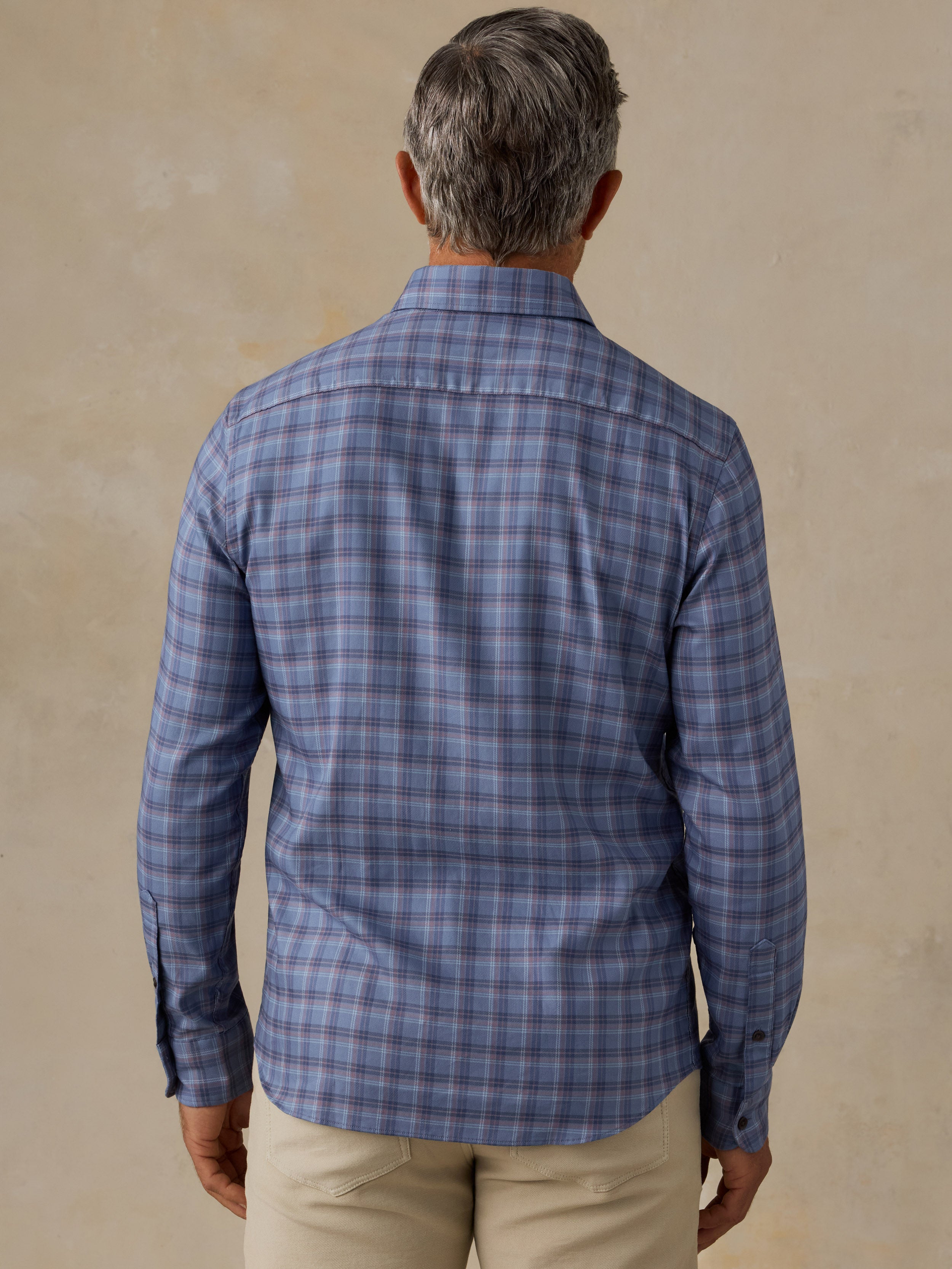 A man viewed from behind wearing a blue gray plaid long-sleeve button-down shirt and khaki pants, standing against a textured beige wall.