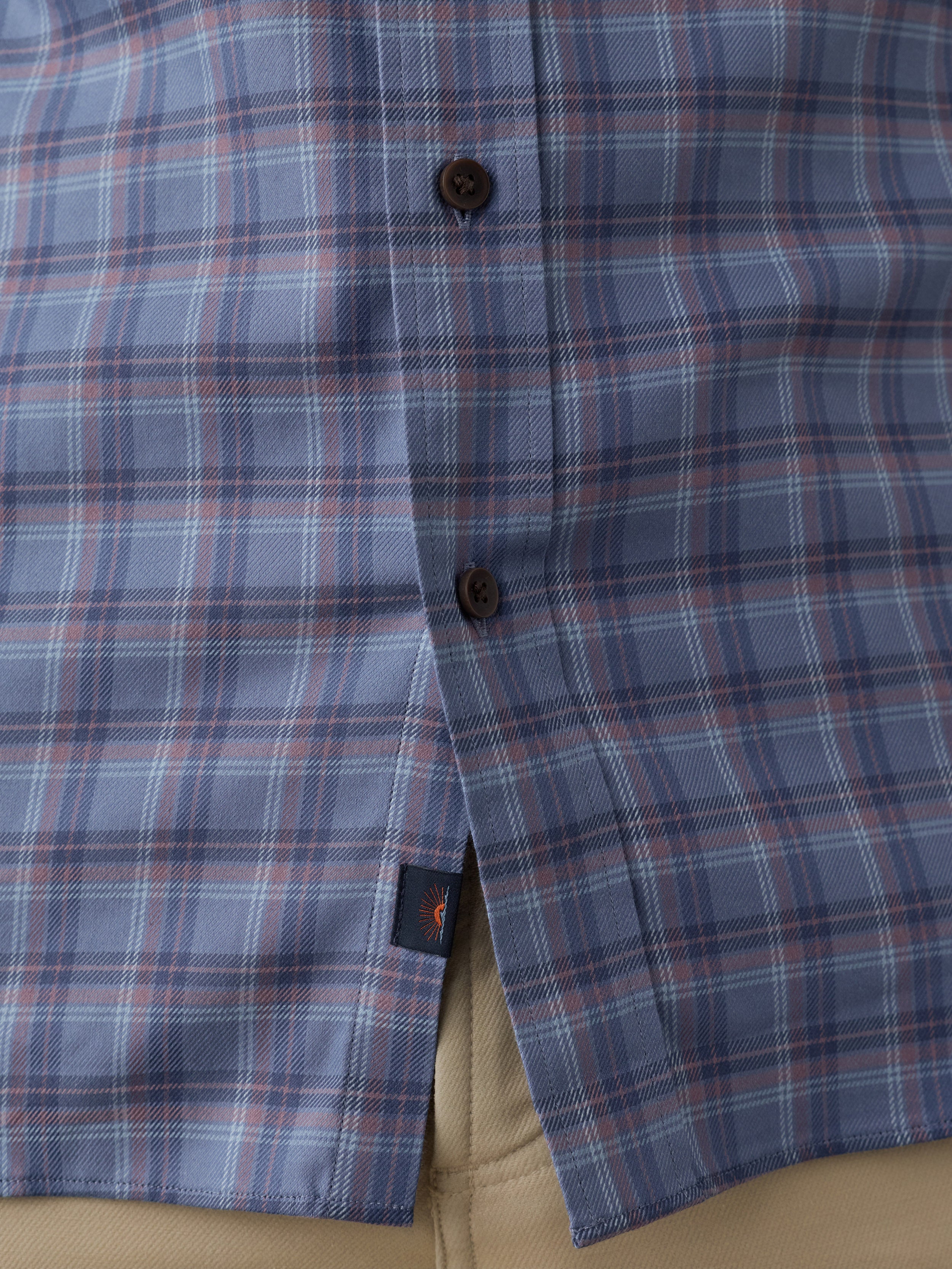 Close-up of a plaid button-down shirt in blue, red, and white, with dark brown buttons and a small Faherty brand tag near the hem.