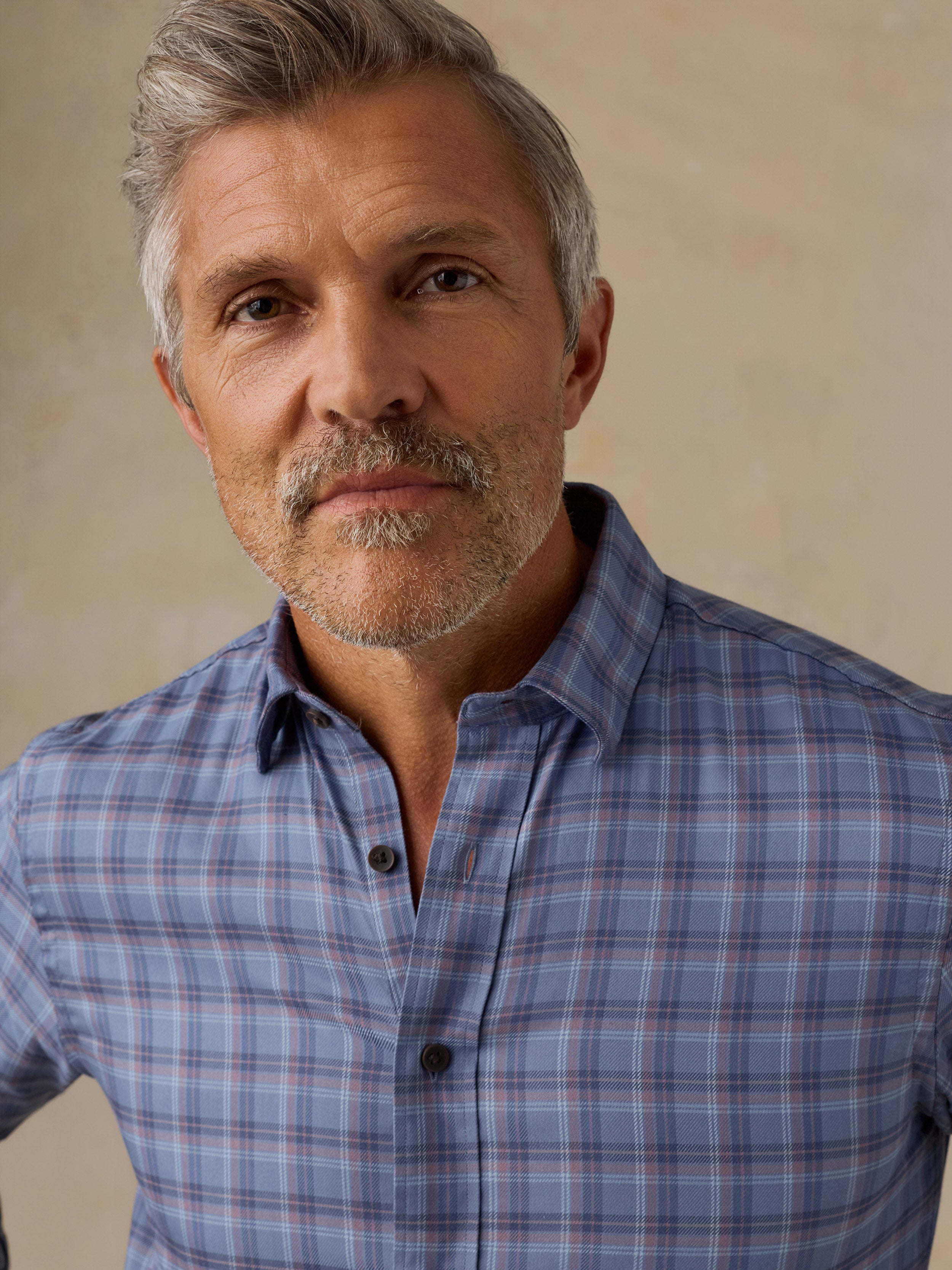 A close-up portrait of a man with gray hair and a beard wearing a blue gray plaid button-down shirt against a textured beige background.