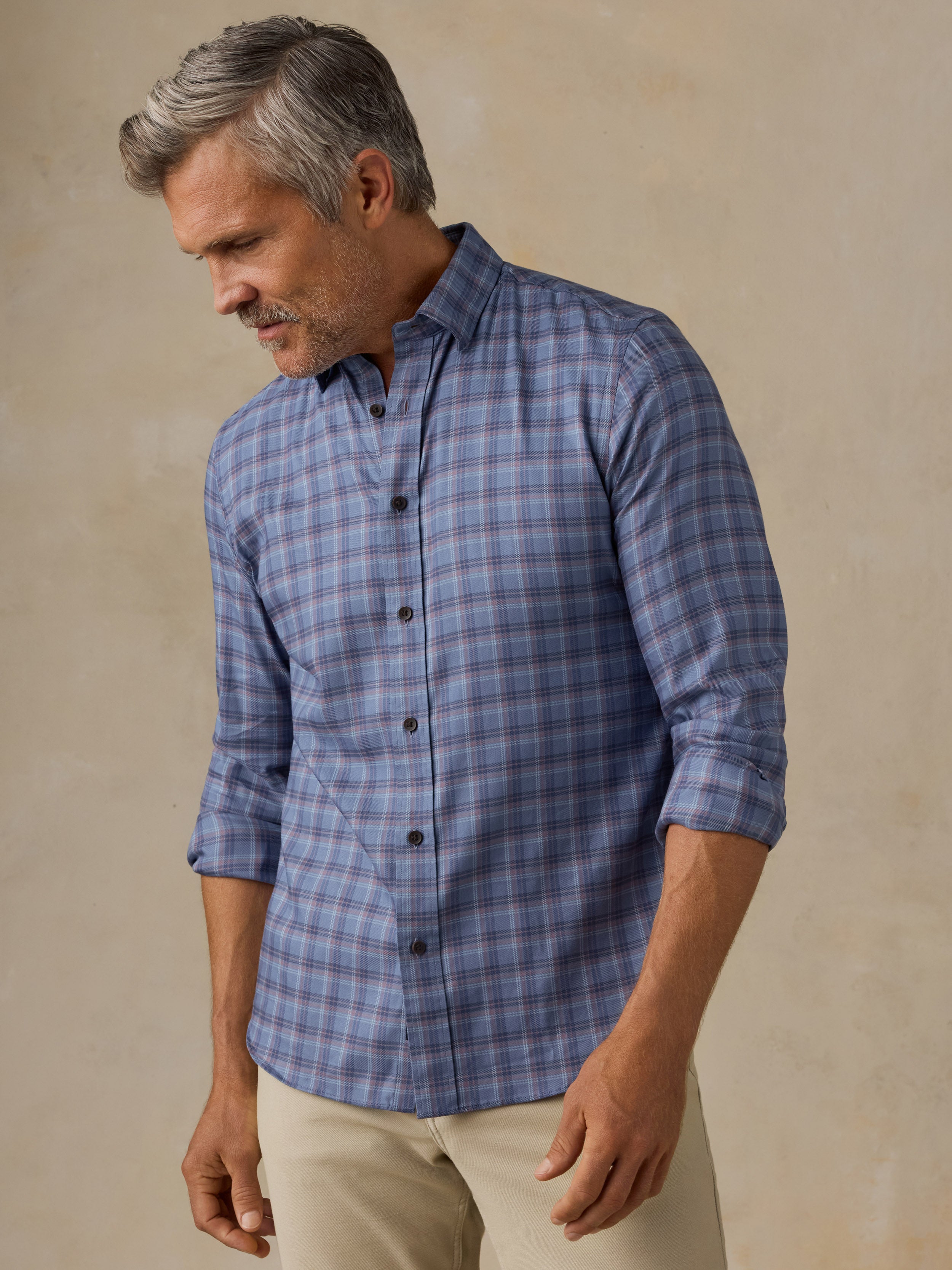 A man wearing a blue gray plaid button-down shirt with beige pants, standing against a textured beige background.