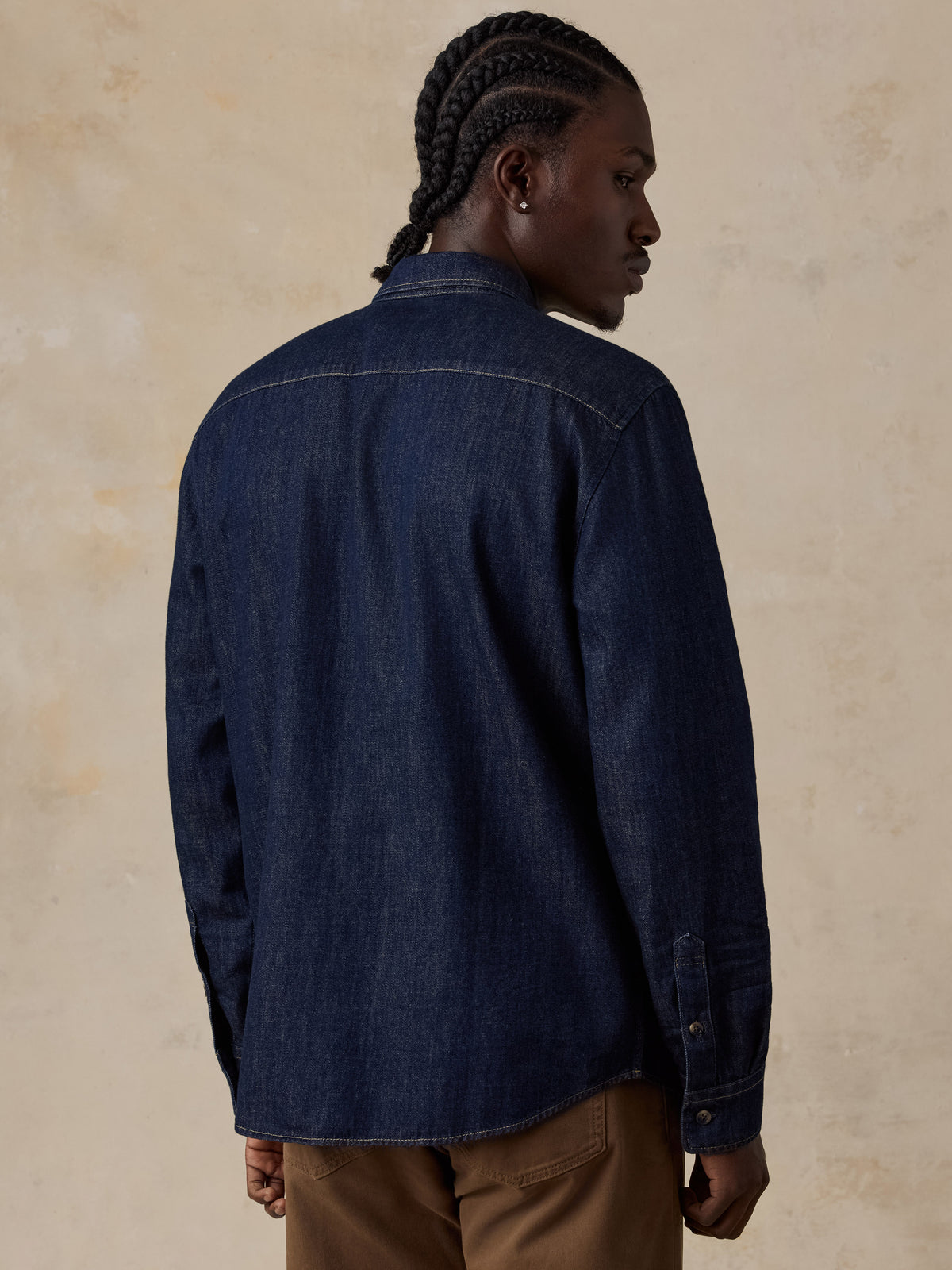 A man shown from the back wearing a dark indigo denim shirt jacket and brown trousers, standing against a textured beige studio background.