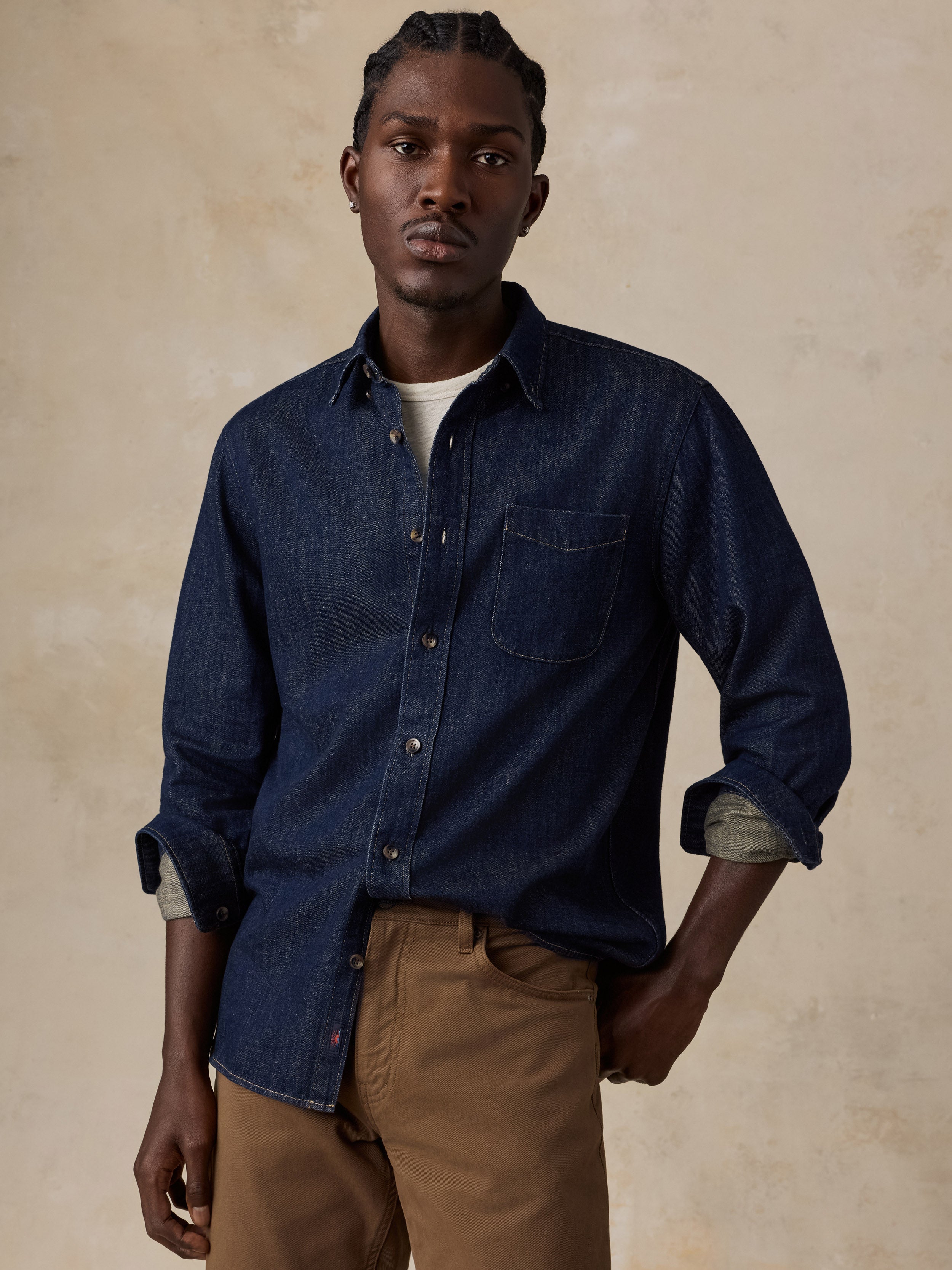 A man wearing a dark indigo denim long-sleeve button-up shirt with rolled-up sleeves, paired with brown trousers, standing against a textured beige background.