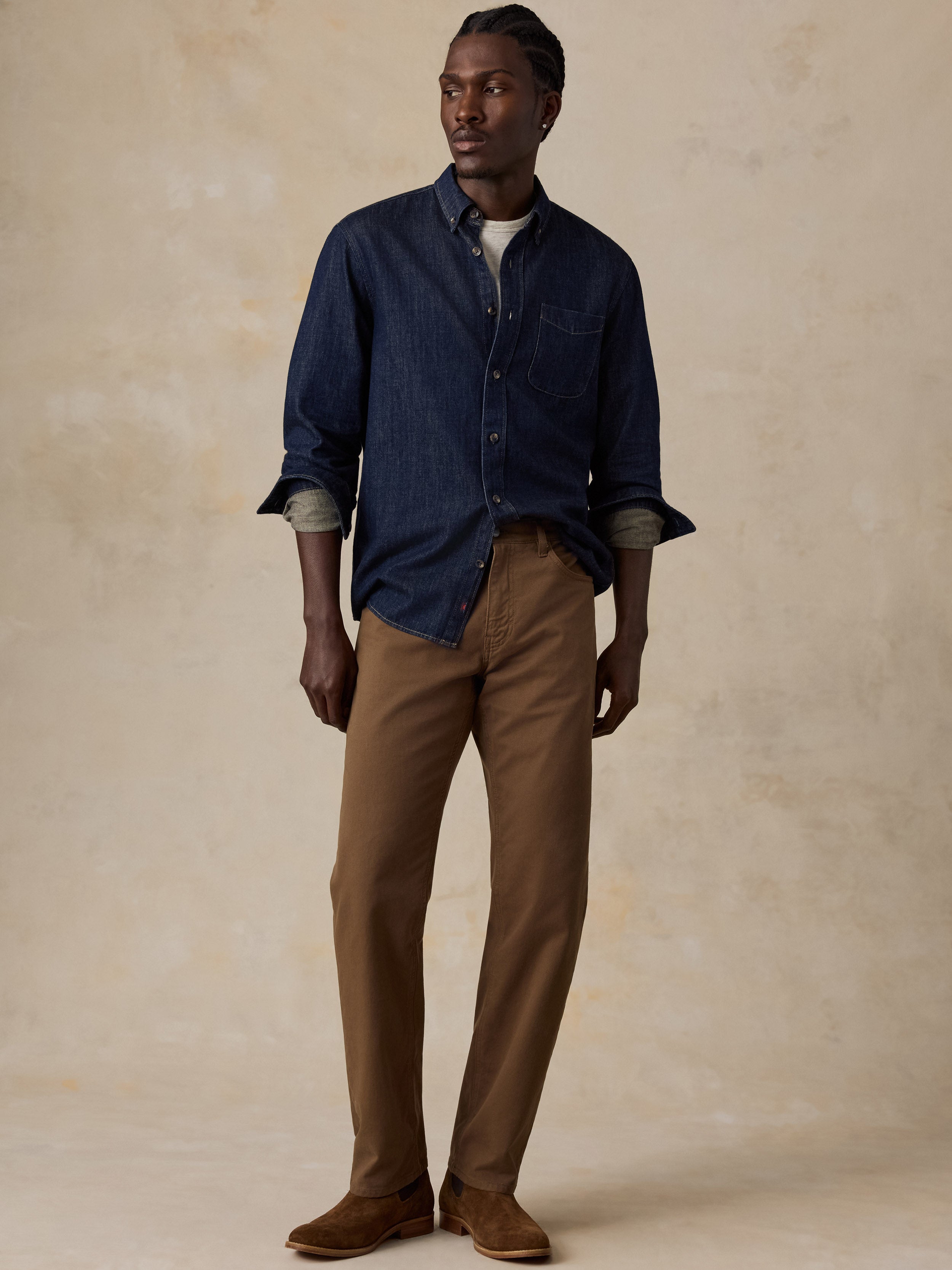 A man modeling a dark blue denim button-down shirt with rolled sleeves, paired with brown trousers and brown suede shoes, standing against a textured beige studio backdrop.