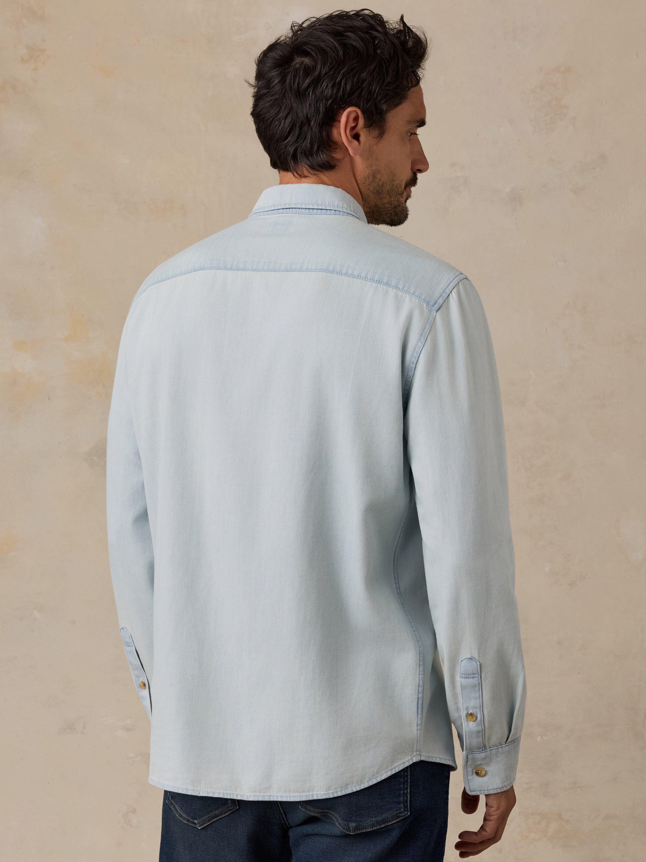 A man shown from the back wearing a light blue long-sleeve button-up shirt and dark blue jeans, standing against a textured beige wall.
