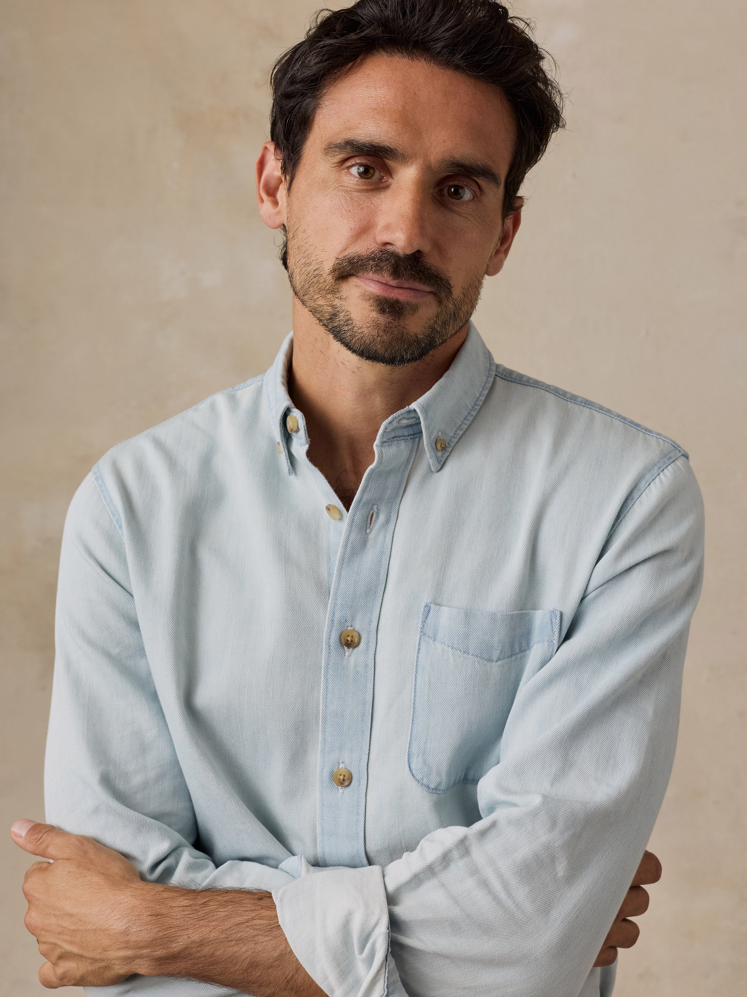 A man wearing a light blue long-sleeve button-down shirt with buttons, posed against a neutral beige textured background.