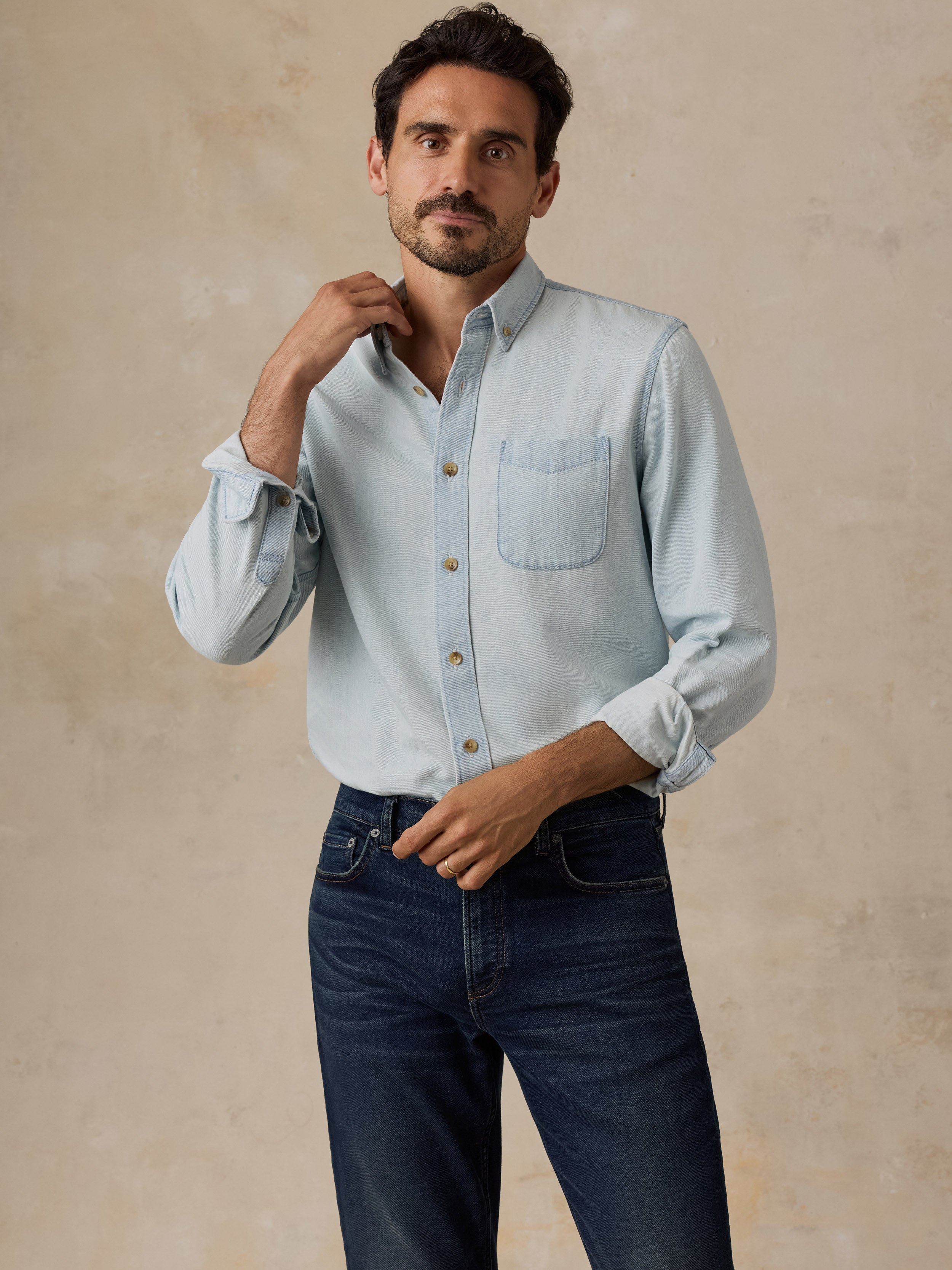 A man wearing a light blue long-sleeve button-down shirt with a chest pocket and dark blue jeans, standing against a textured beige studio backdrop.