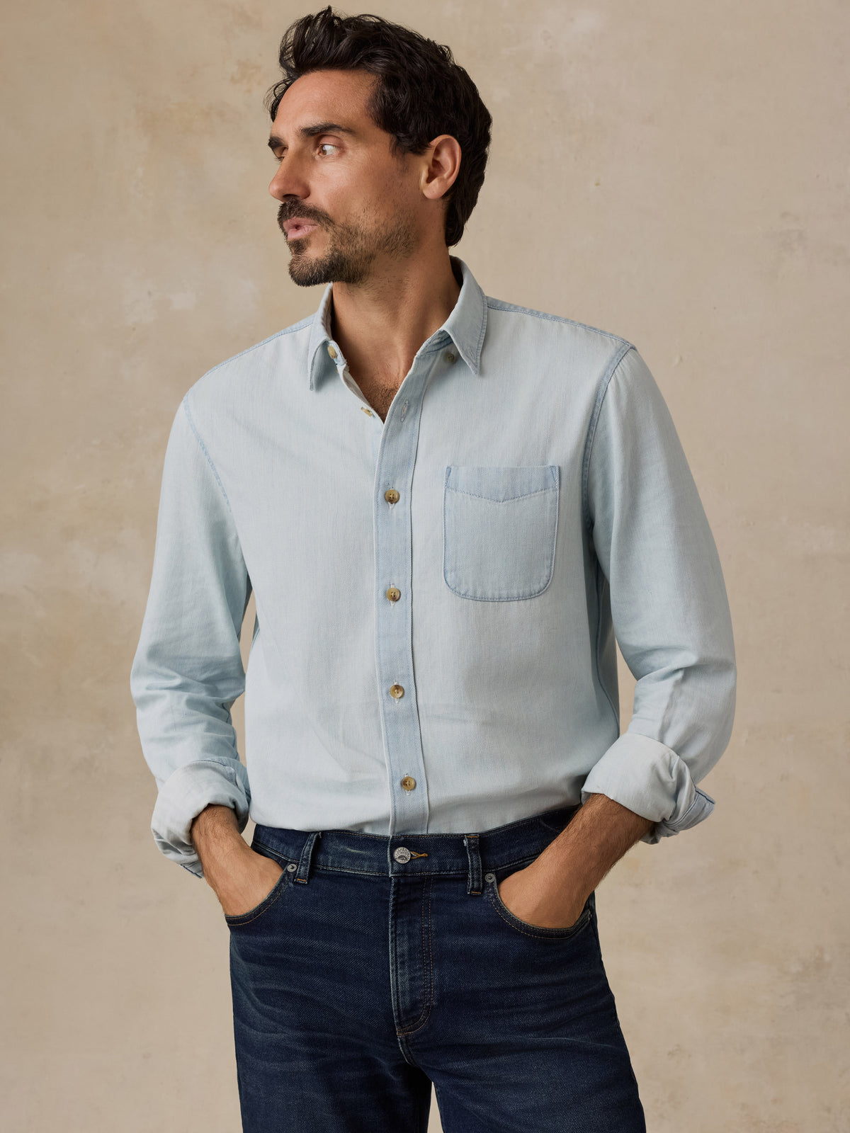 A man wearing a light blue long-sleeve button-down shirt with rolled-up sleeves and dark blue jeans, standing against a textured beige background.