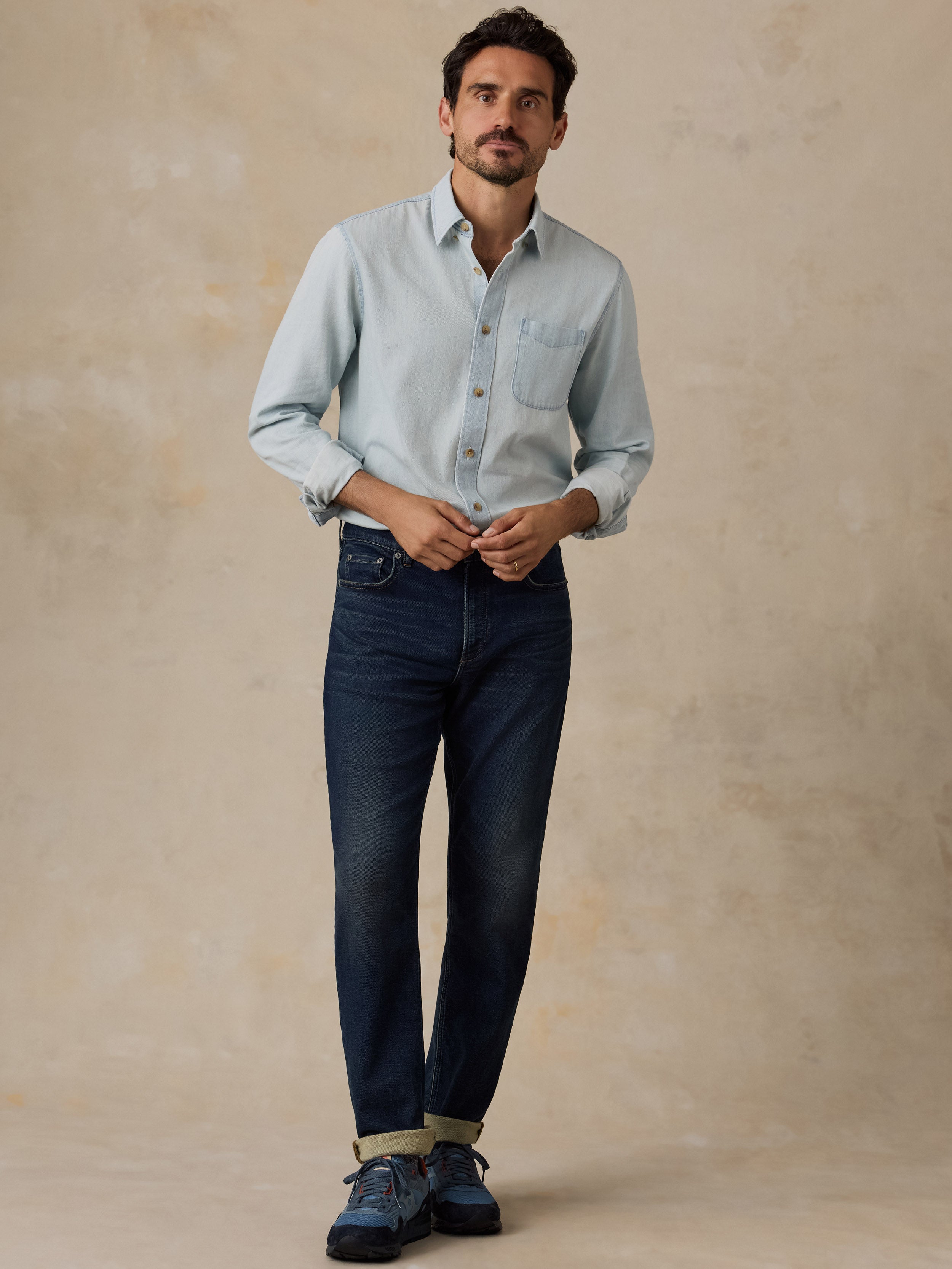 A man wearing a light blue long-sleeve button-down shirt, dark blue jeans, and blue sneakers, standing against a textured beige studio backdrop.