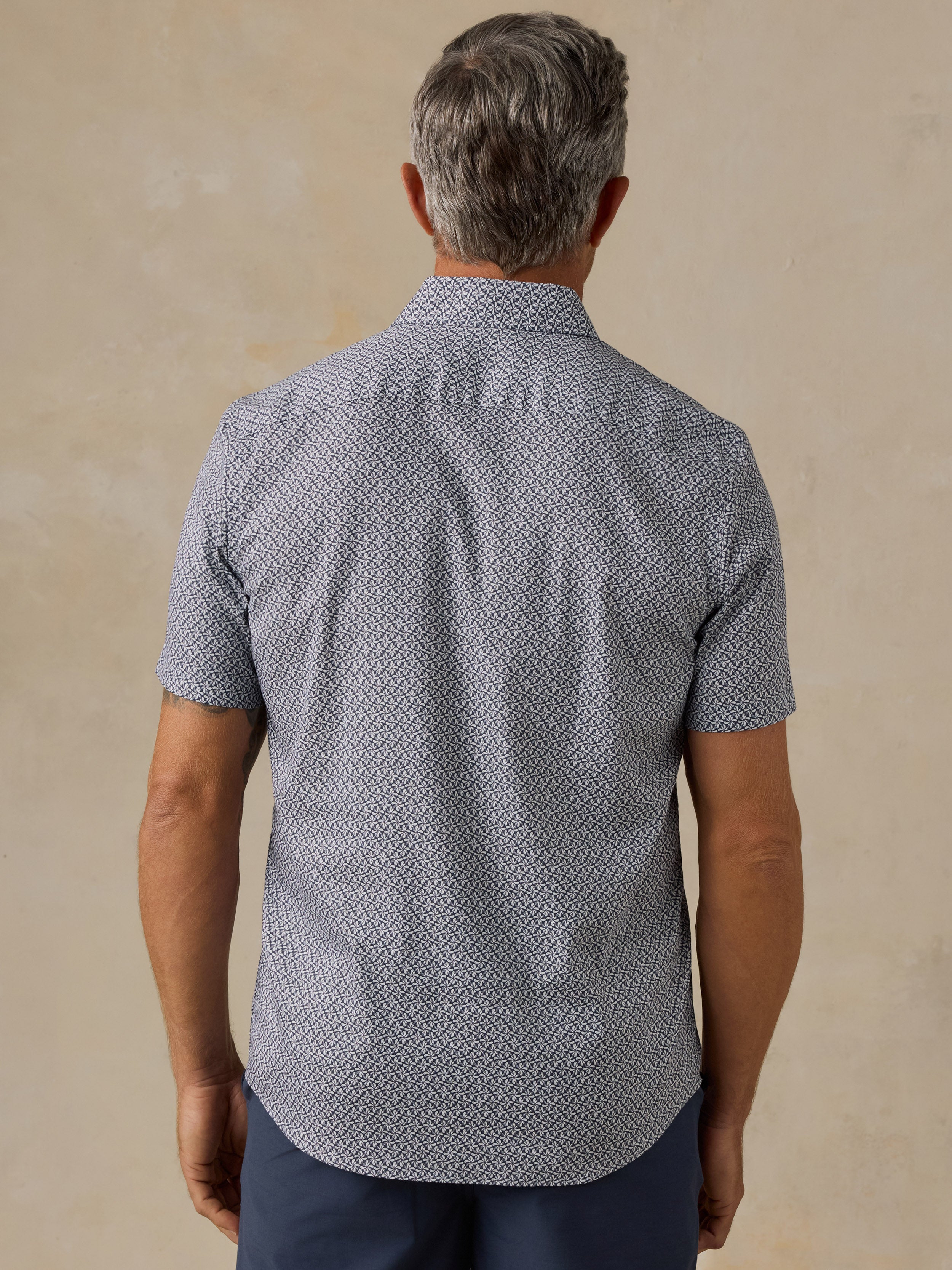 A man viewed from behind wearing a short-sleeve button-up shirt with a dense small-scale geometric pattern in light gray and white, paired with navy blue trousers, set against a textured beige background.