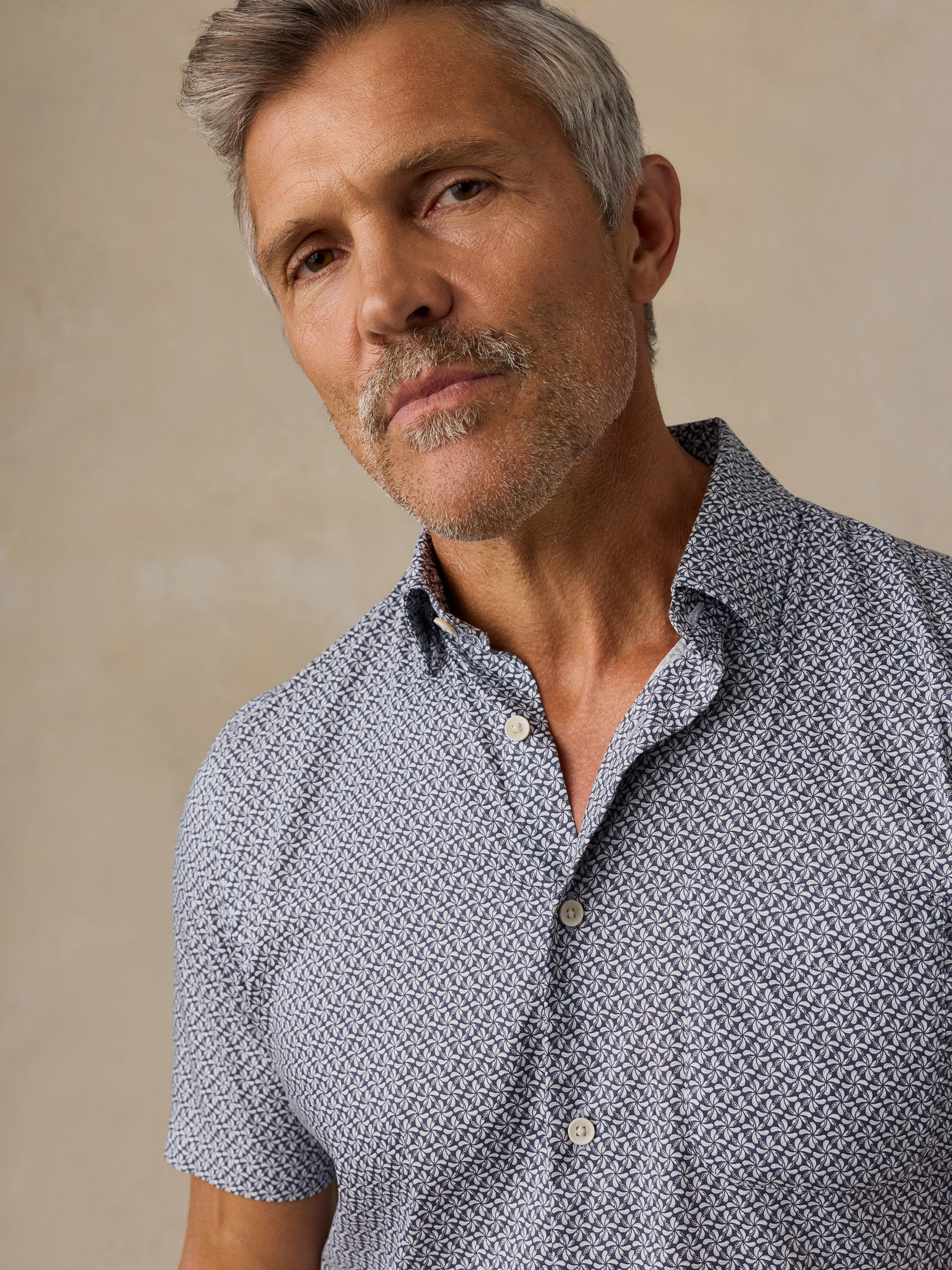 A close-up shot of a man wearing a short-sleeve button-down shirt with a blue and white geometric pattern, set against a neutral beige background.