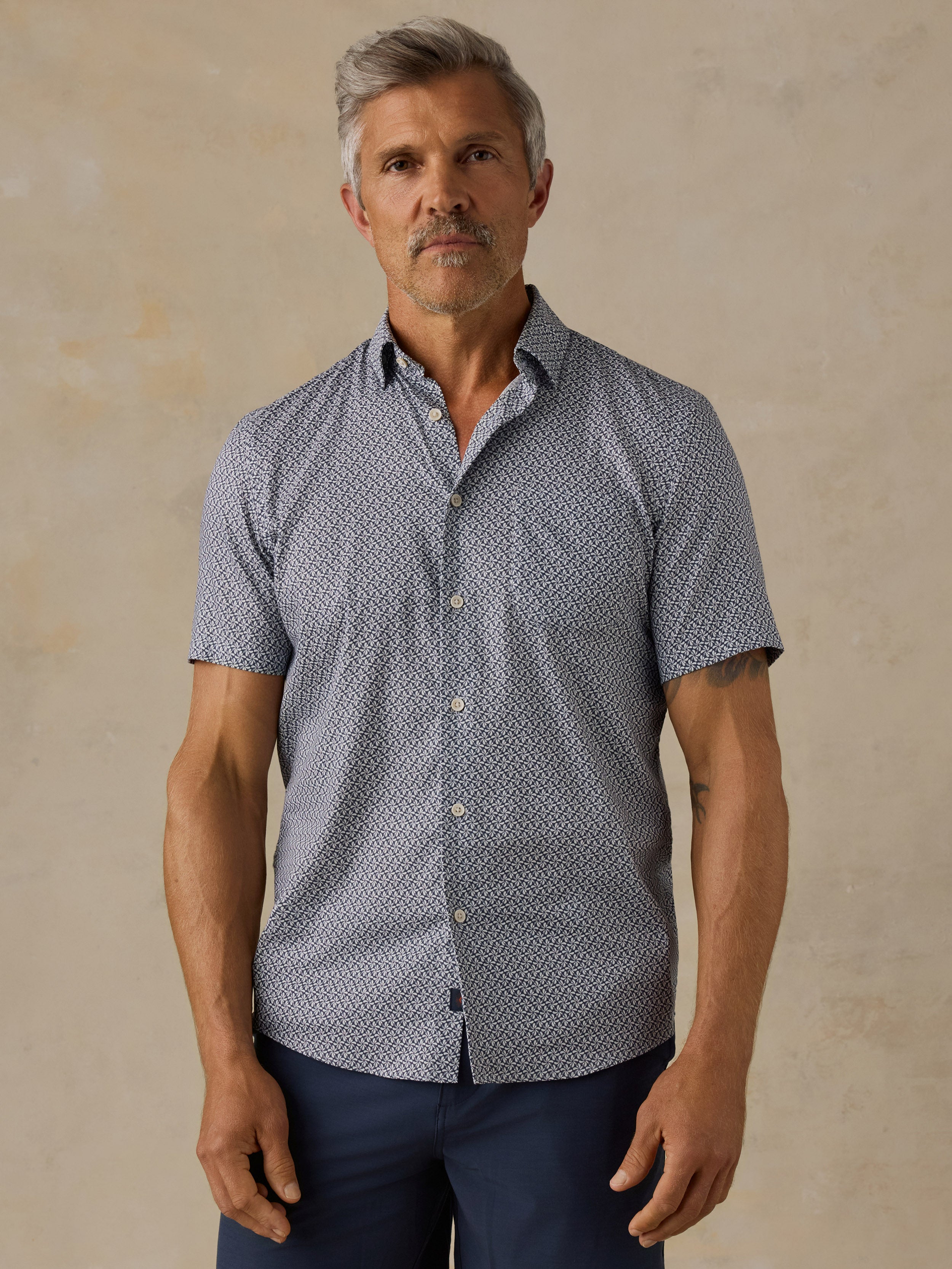 A man wearing a short-sleeve button-up shirt with a small blue and white geometric pattern, paired with navy pants, standing against a textured beige background.