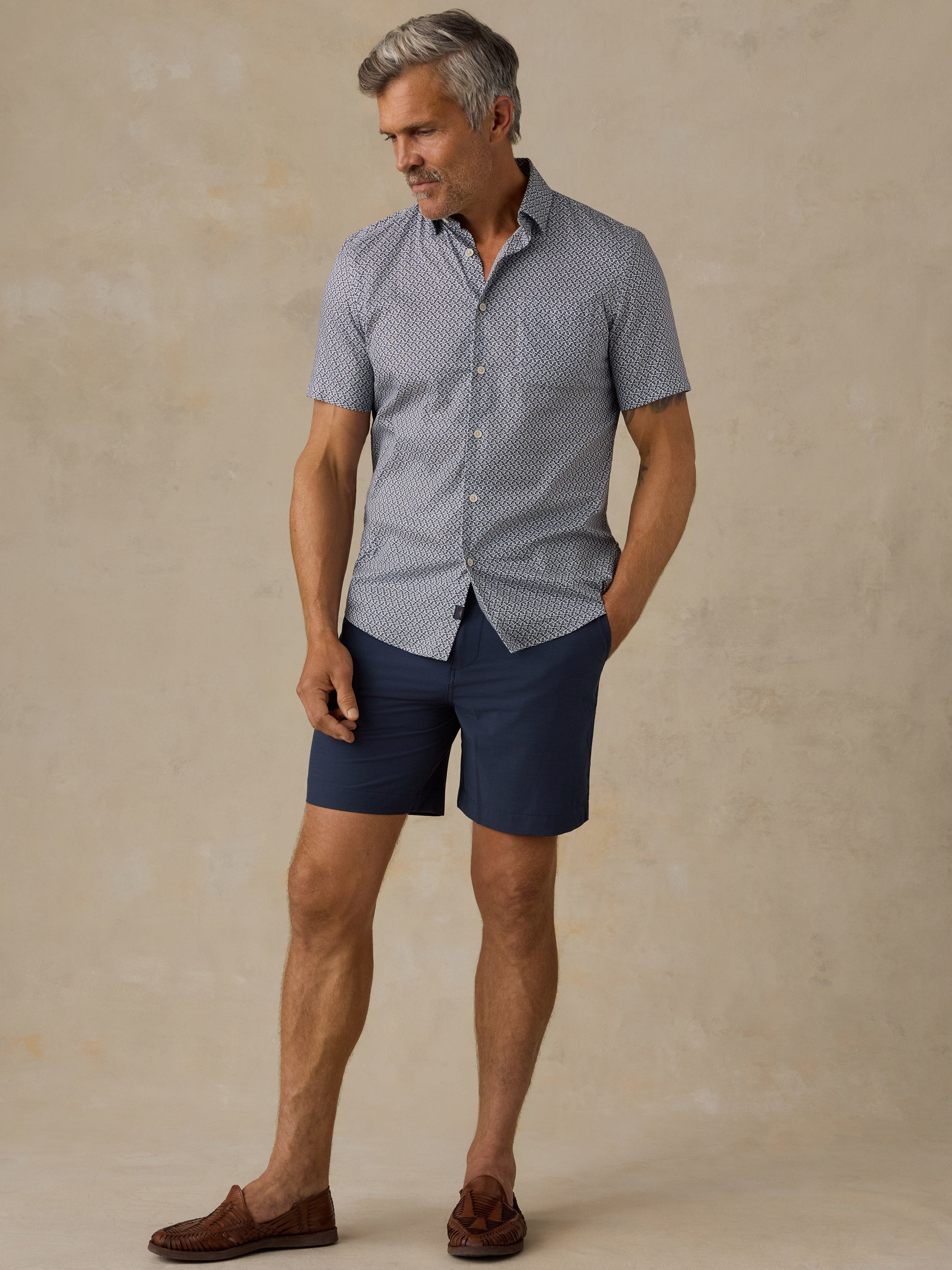 A man modeling a short-sleeve button-up shirt with a small blue and white geometric pattern, paired with navy blue shorts and brown suede loafers, set against a textured beige studio backdrop.