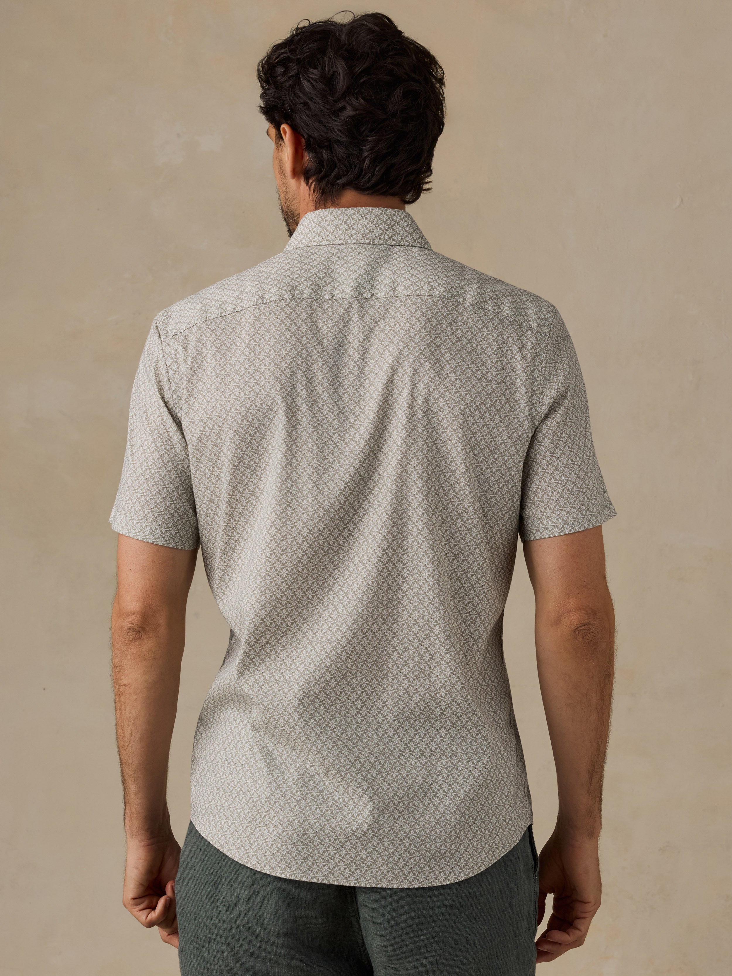 A man viewed from behind wearing a short-sleeve button-up shirt with a light beige and green geometric pattern, paired with dark green shorts, set against a textured beige background.
