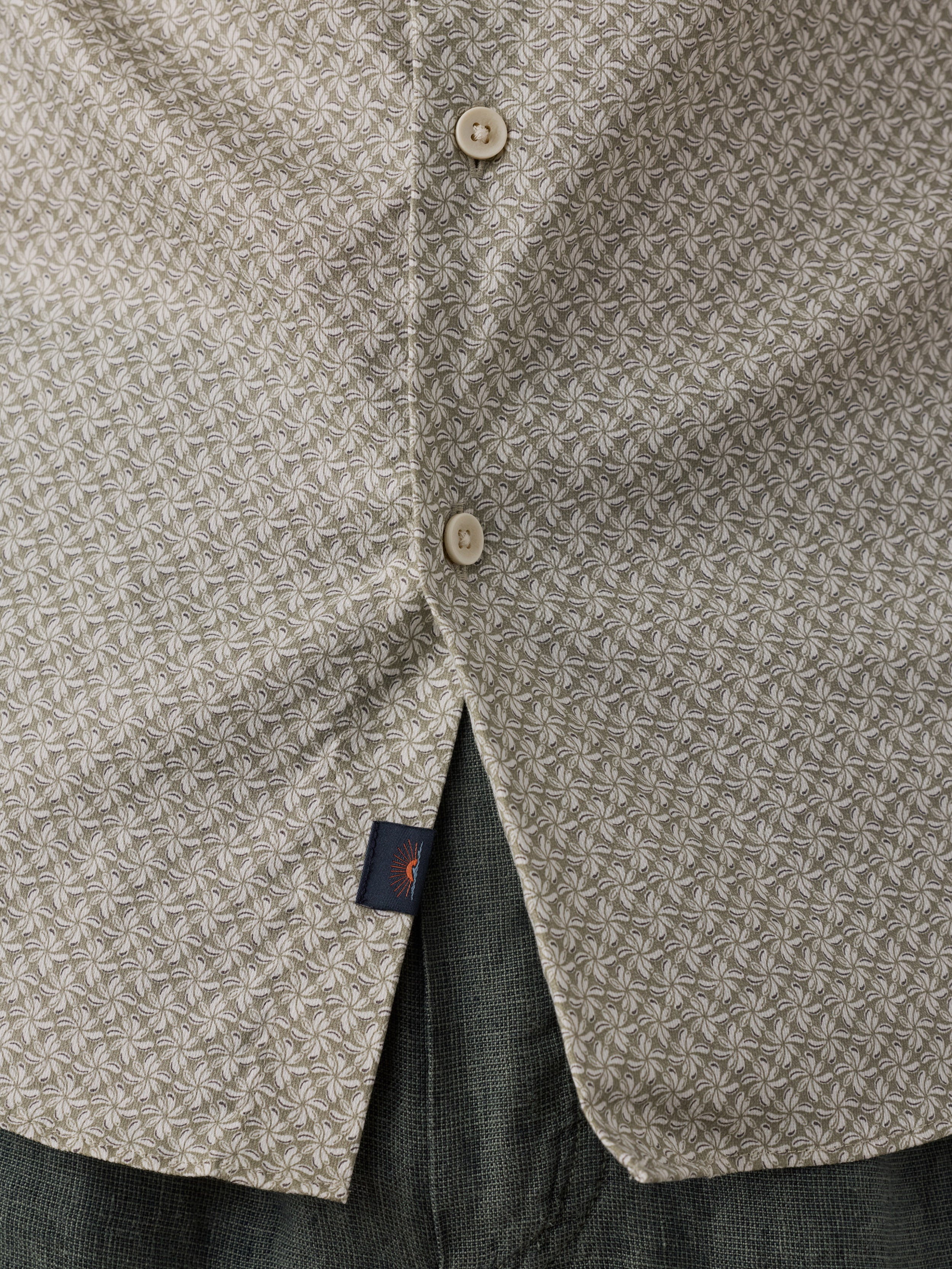 Close-up of a beige and green geometric patterned button-down shirt with a small navy Faherty brand sun and waves tag.