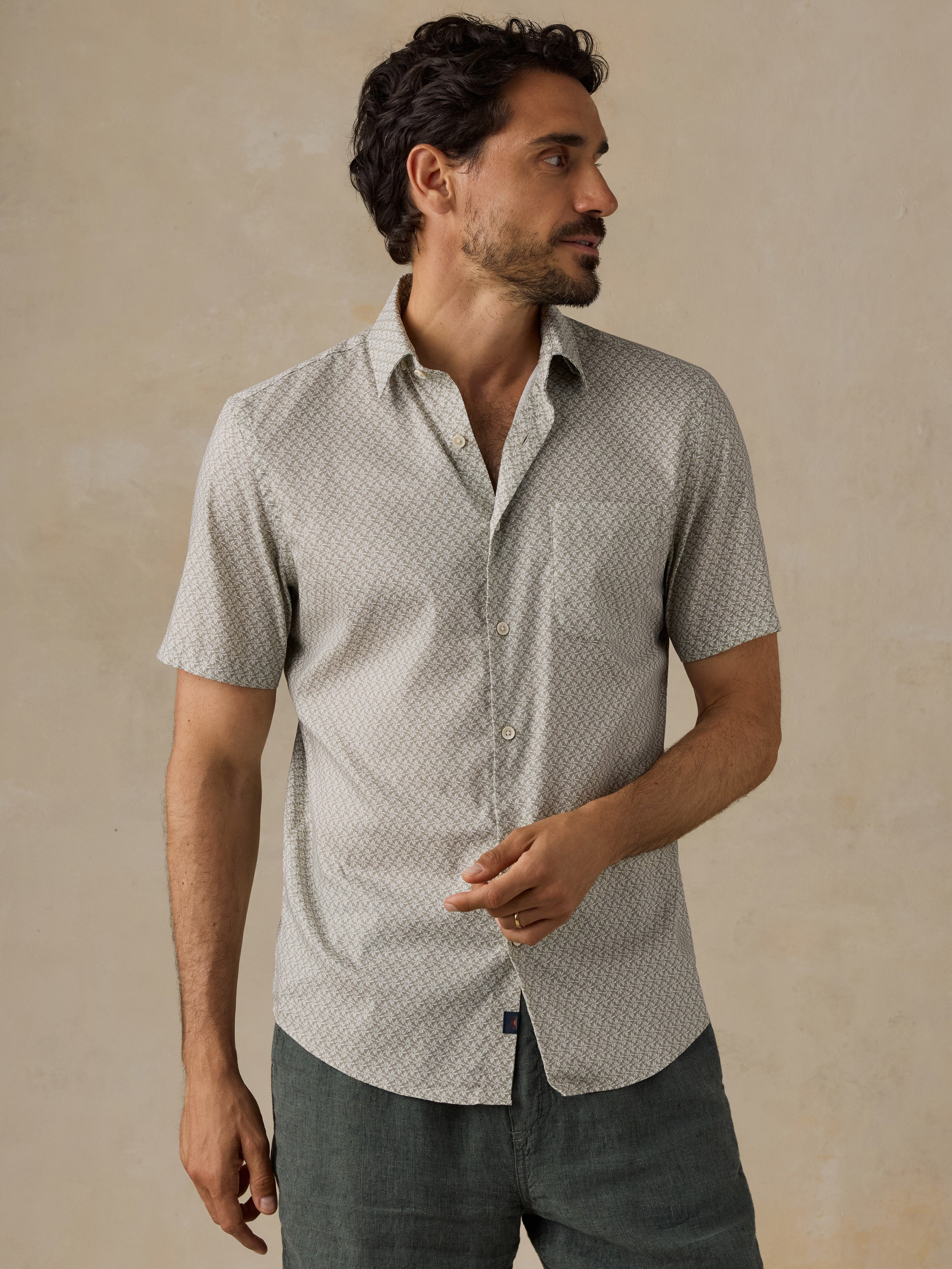 A man wearing a light green short-sleeve button-up shirt with a subtle geometric pattern, paired with dark green shorts, standing against a textured beige background.